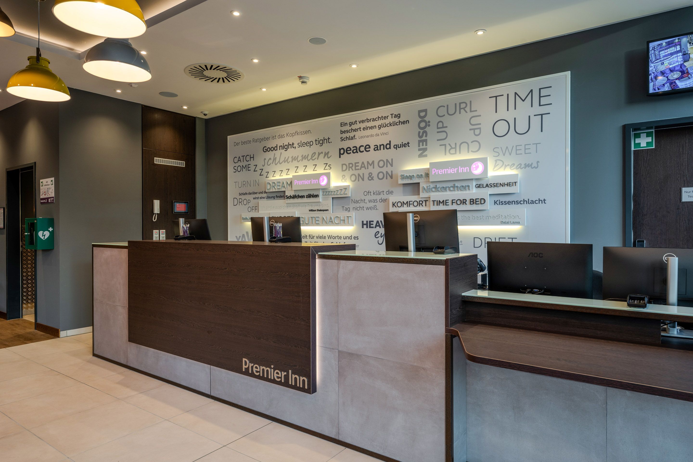 A modern reception area at the Premier Inn Hotel with a stylish design. The wall is decorated with inspiring quotes. Computers and desk lamps are visible.