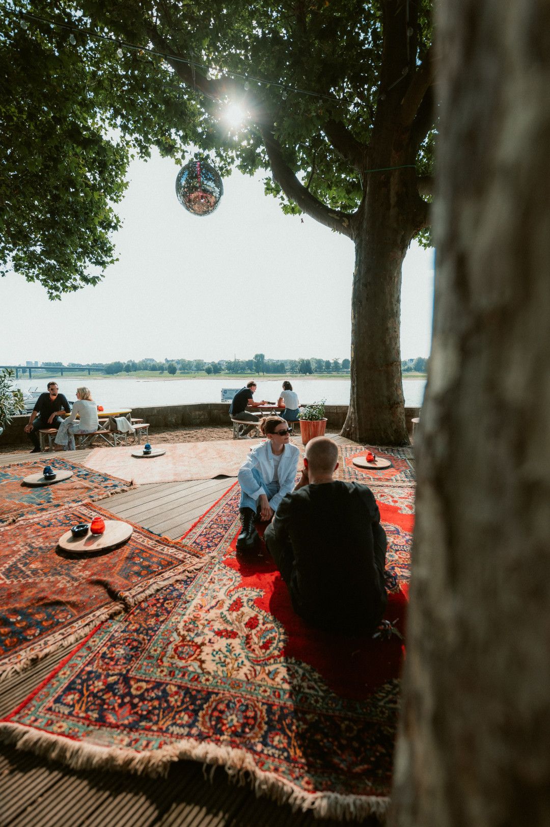 Auf bunten Teppichen sitzen Gäste im Biergarten am Rhein