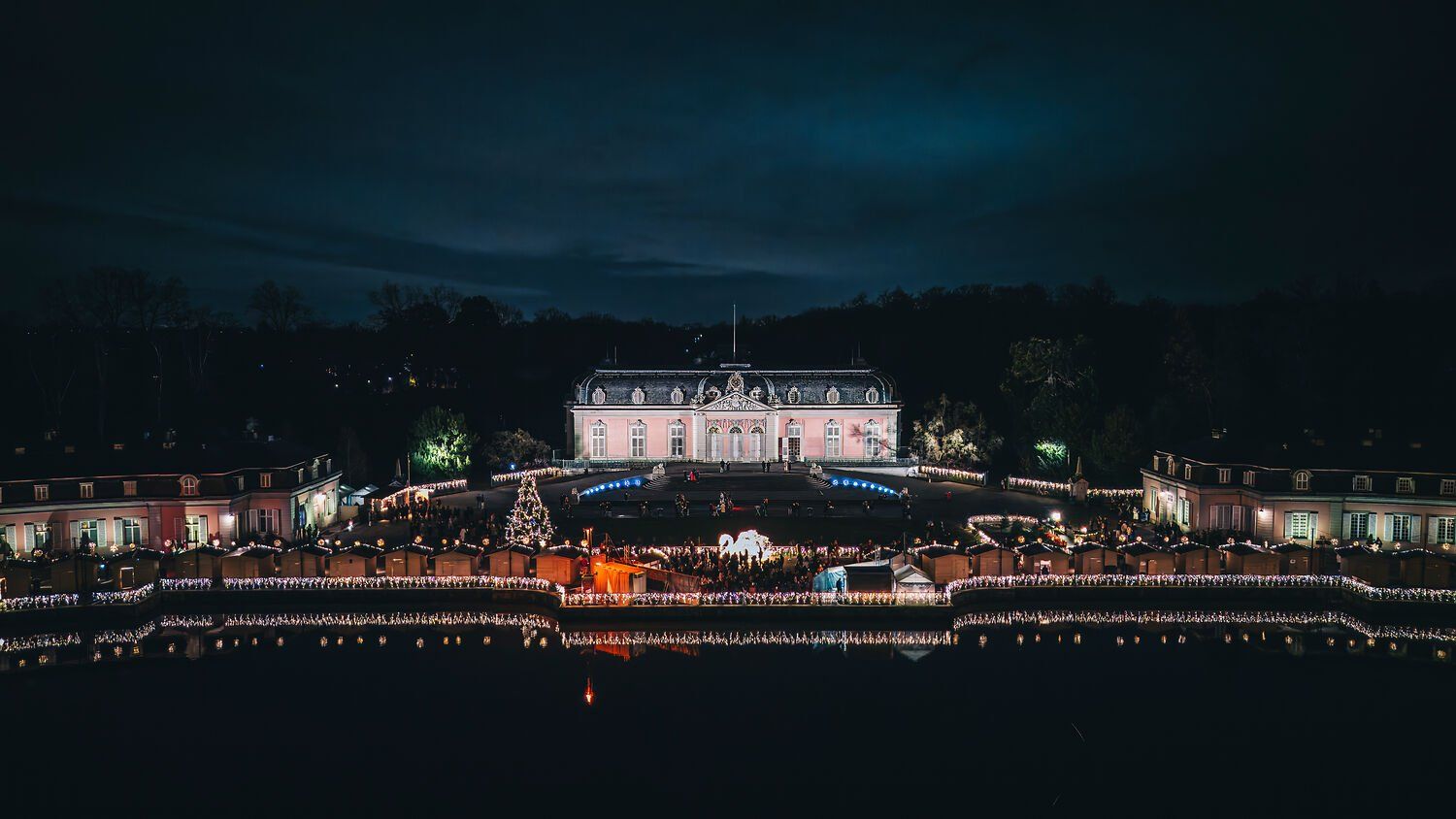 Christmas market at Benrath Palace at night, with illuminated stalls, a Christmas tree, and its reflection in the water.