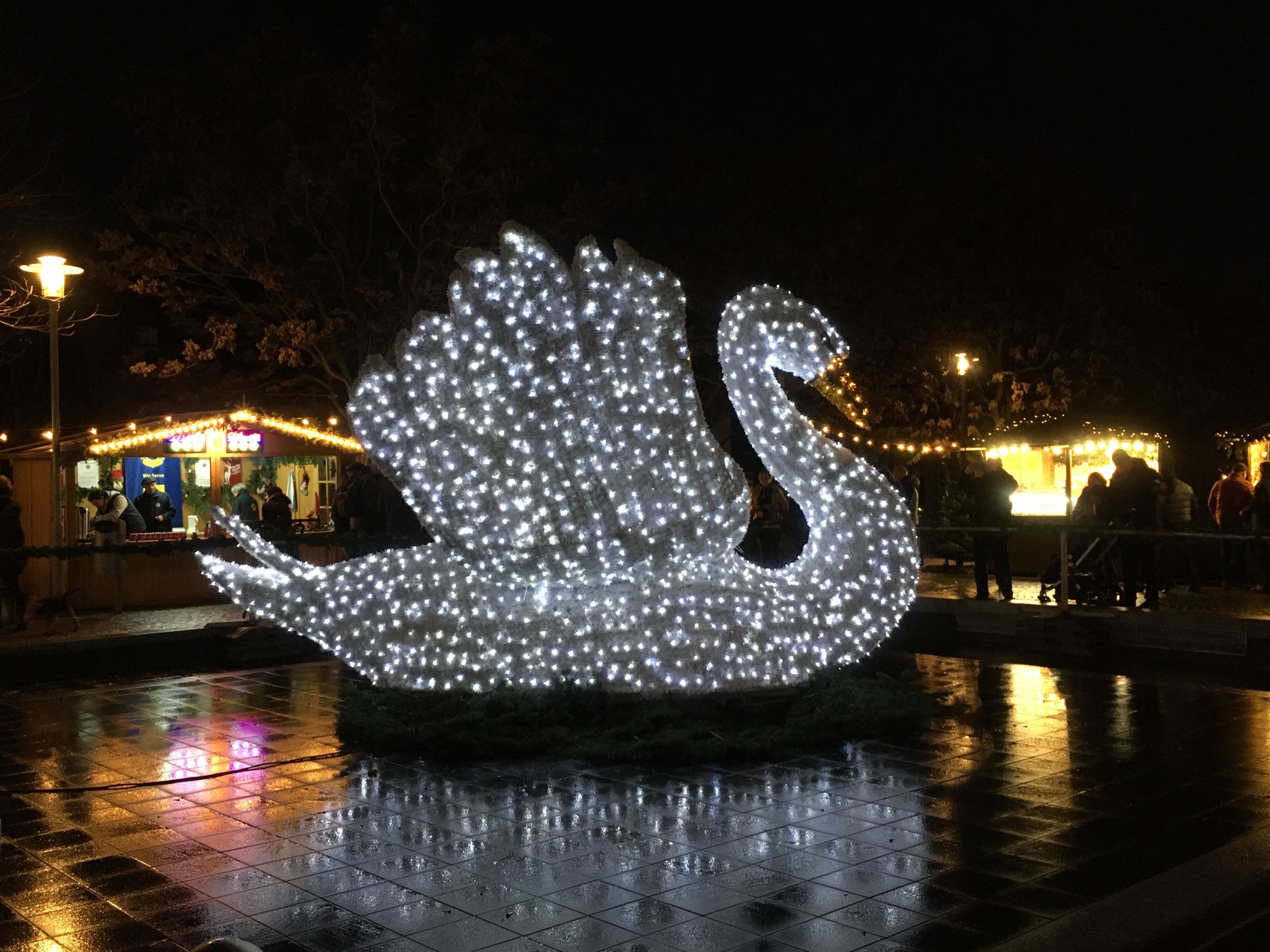 A glowing swan at the Kleve Christmas market at night, with stalls and wet, reflective pavement.