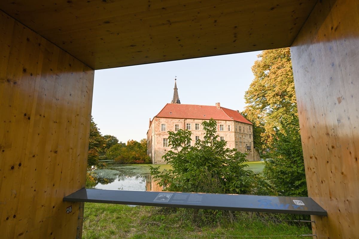 View of Lüdinghausen Castle through a wooden frame, surrounded by trees and a moat.