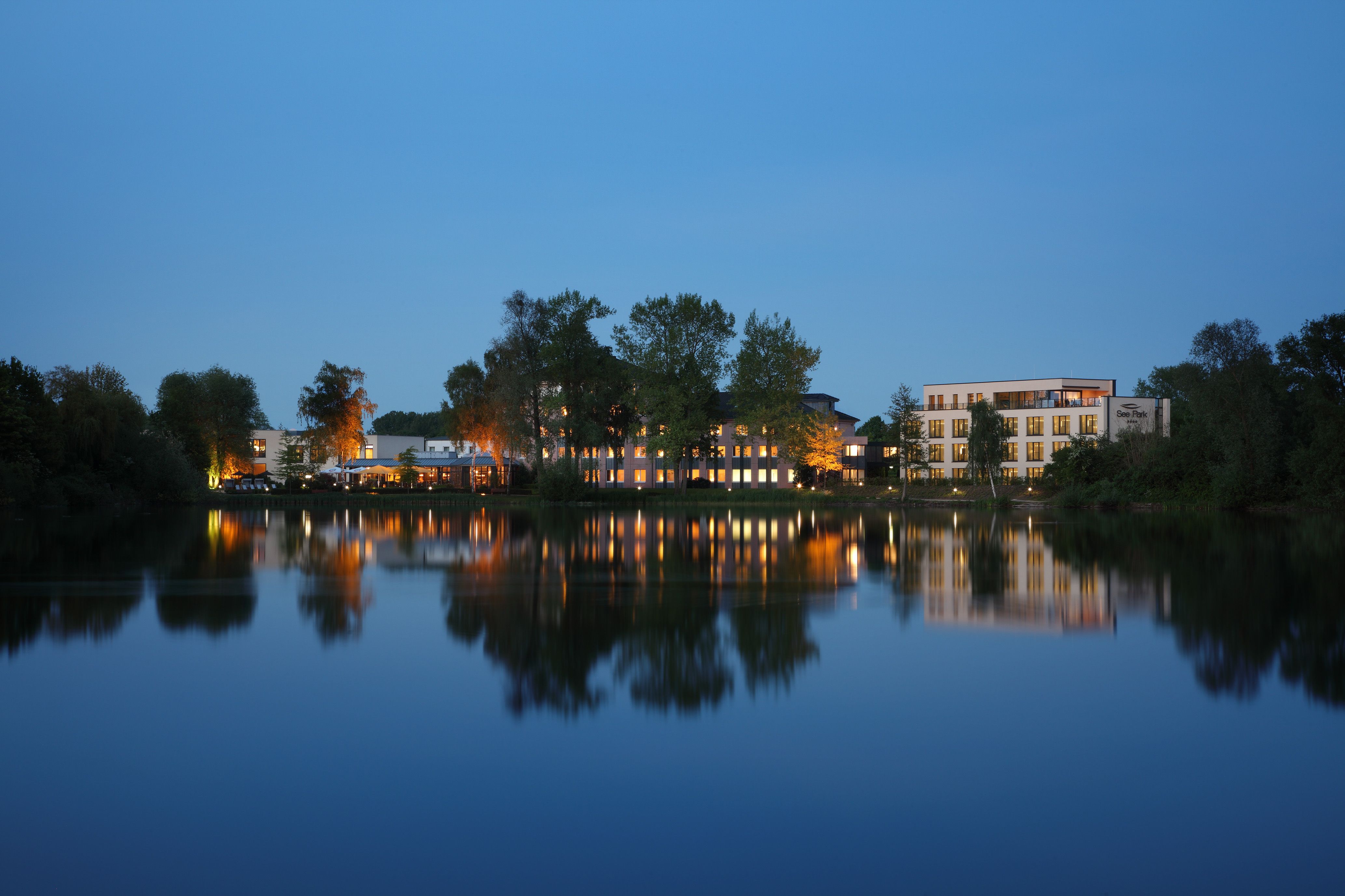 Evening view of an illuminated building by the lake, park with trees and reflection in the water