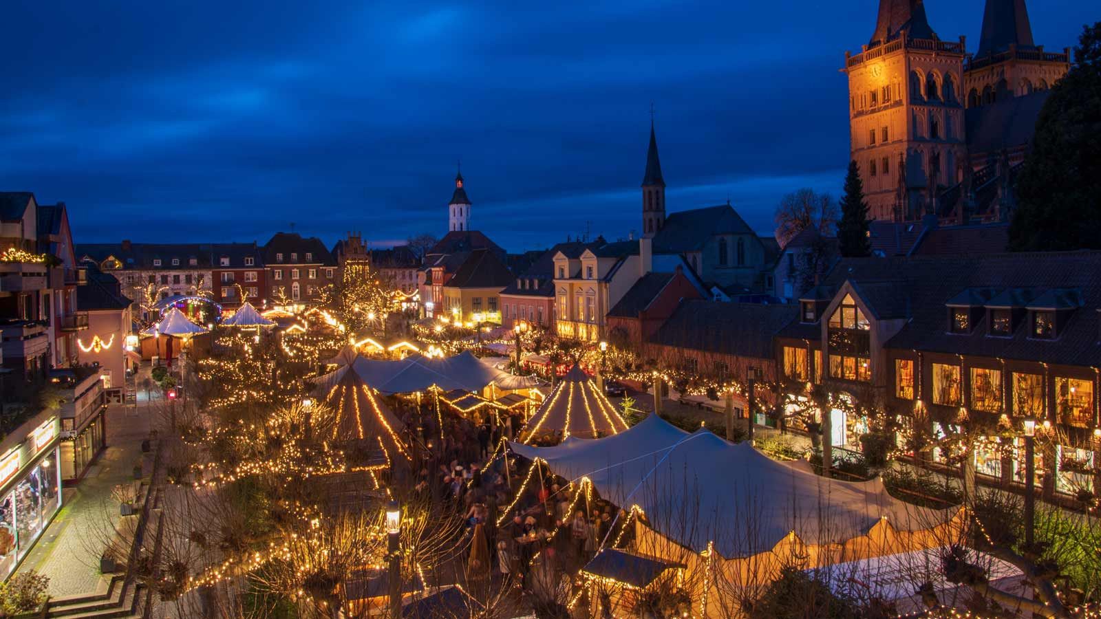 The Christmas market in Xanten shines at night in a festive glow of lights with numerous stalls and tents.
