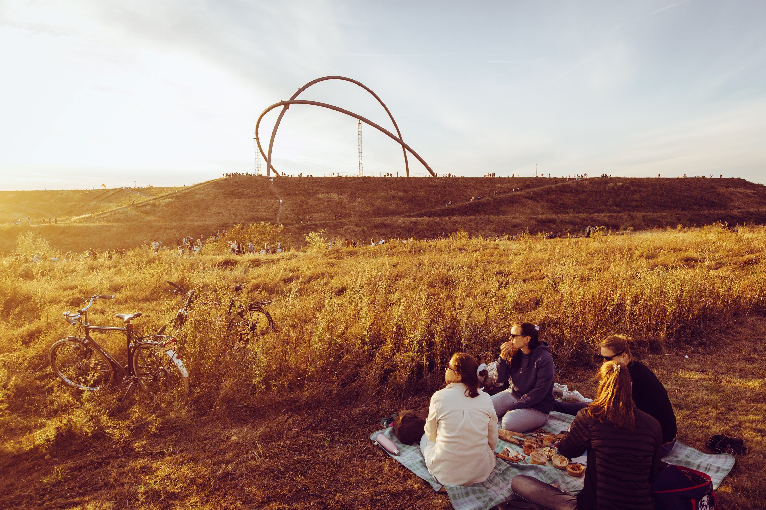 Gruppe picknickt auf Decke im Sonnenuntergang, im Hintergrund ist Skulptur "Observatorium" auf Halde zu sehen 