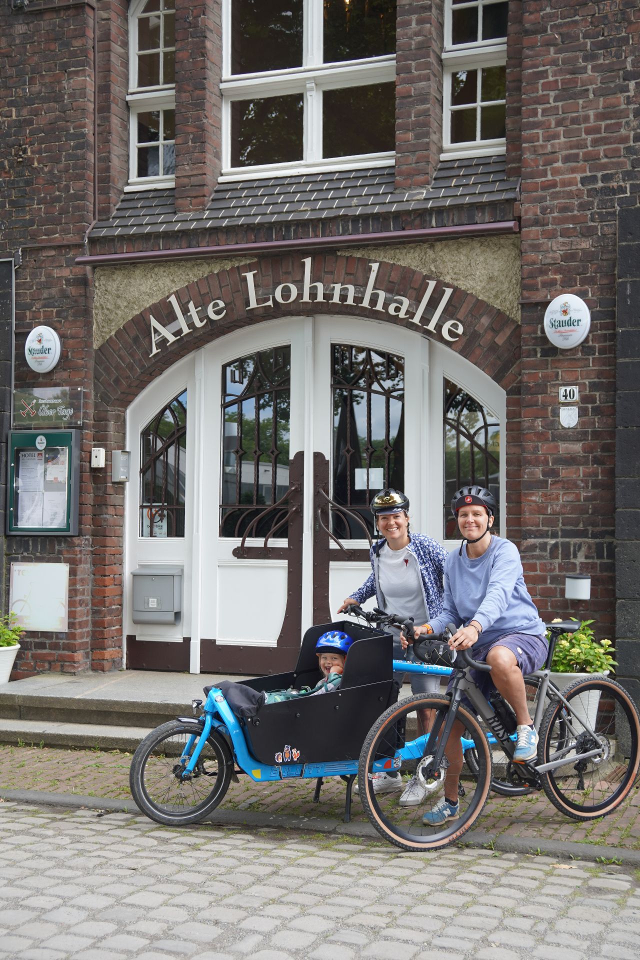A family of three on bicycles stands in front of the Alte Lohnhalle hotel