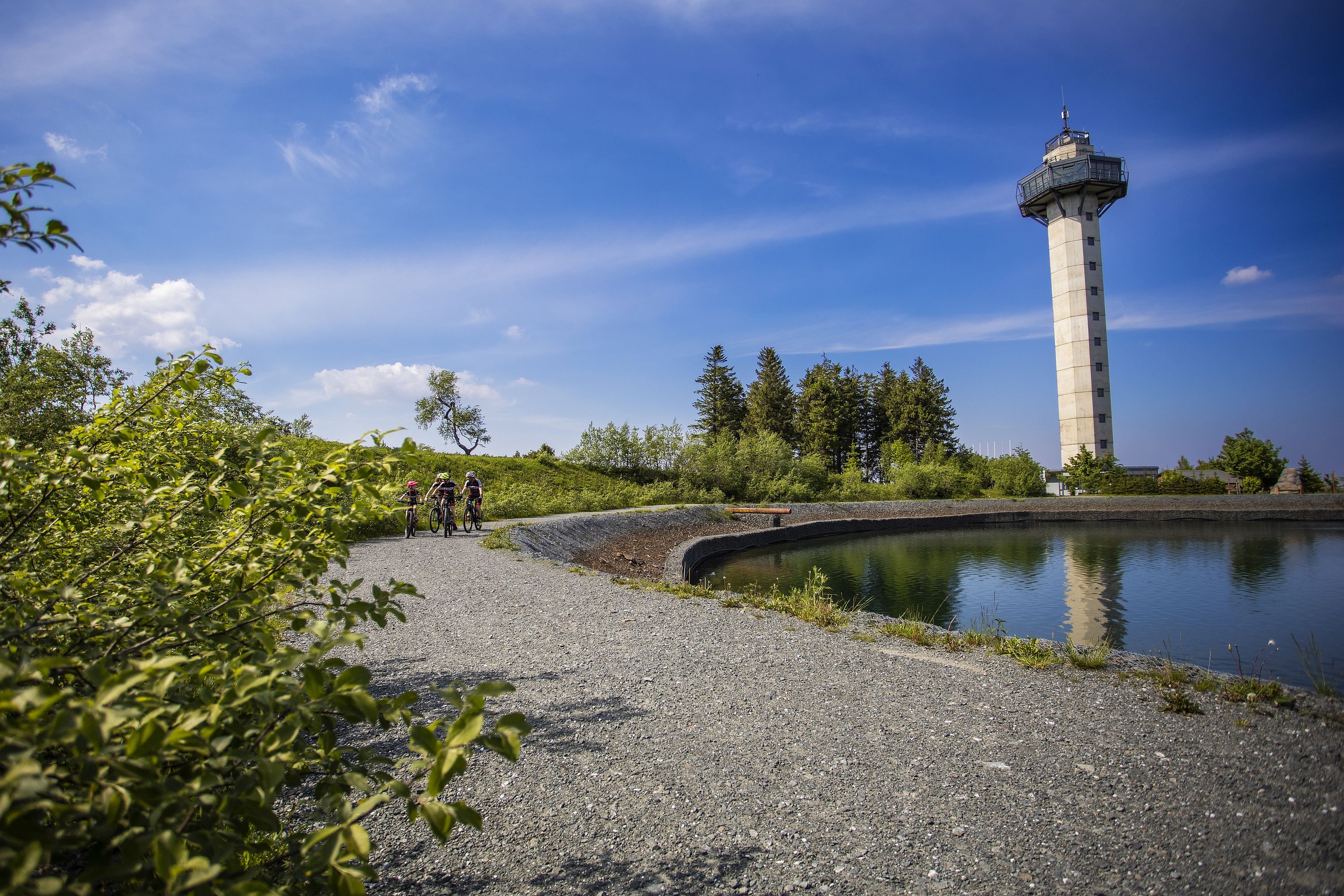 Cyclists round a mountain lake. The Hochheideturm tower is in the background.