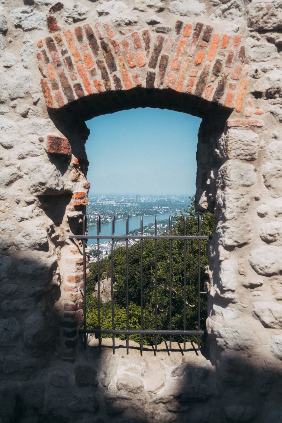 An old stone window with bars offers a picturesque view of the Rhine and the surrounding countryside. The sky is clear and blue.