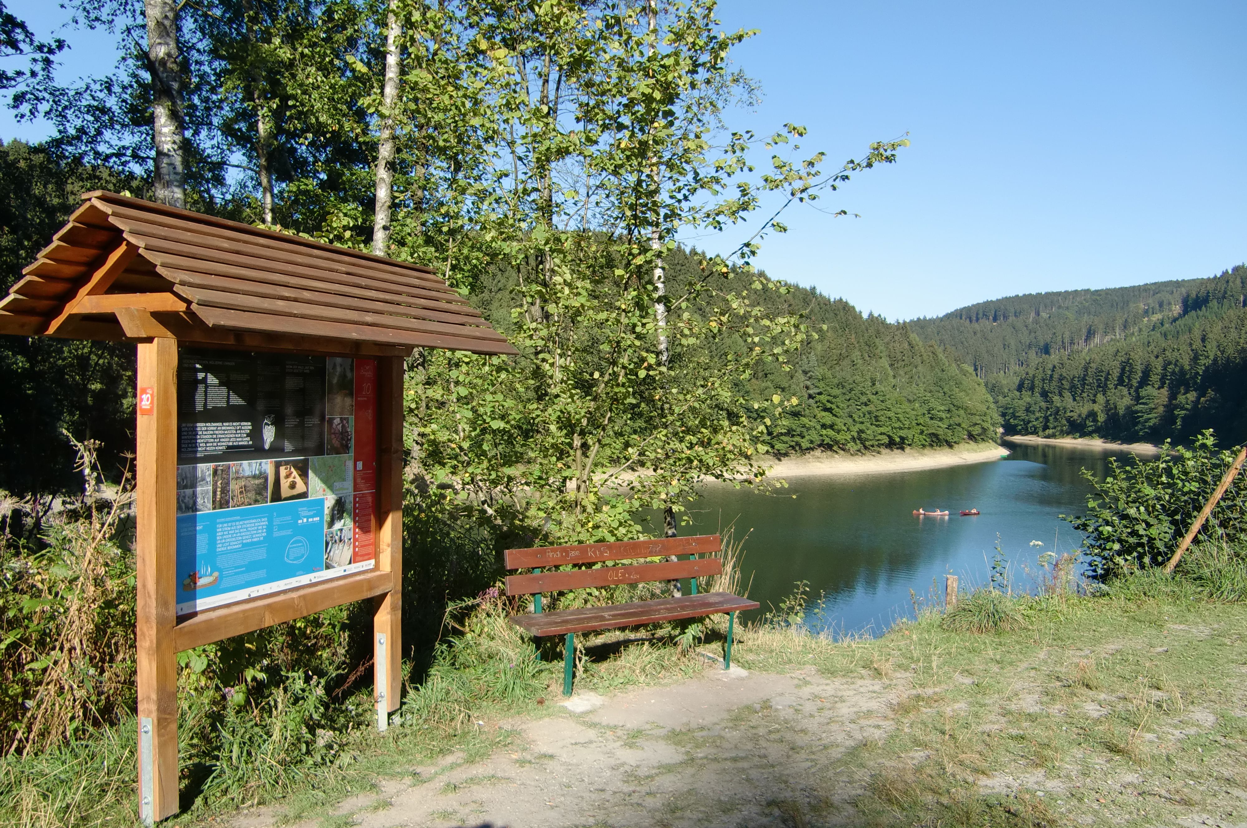An information sign and a bench on the shore of the Aggertal Reservoir, with wooded hills in the background.