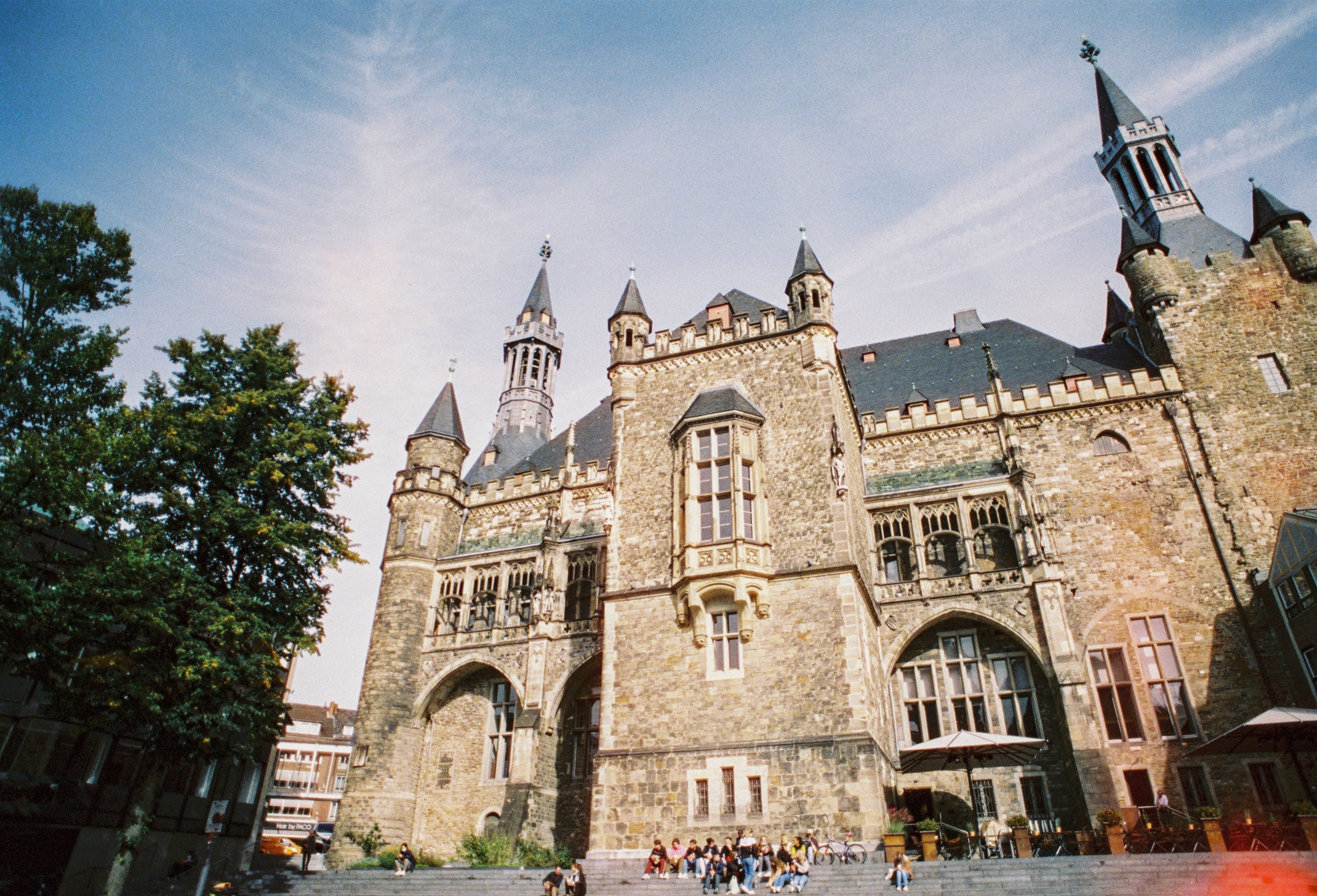 Aachen City Hall, with its striking towers and Gothic elements, surrounded by trees and people on the steps in front of it.