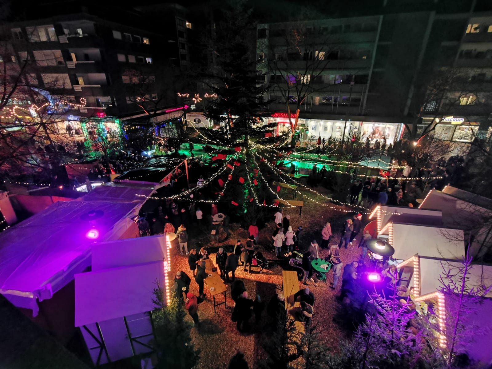 Aerial view of the Christmas market in Gronau at night. People stroll between festively illuminated stalls and a large Christmas tree.