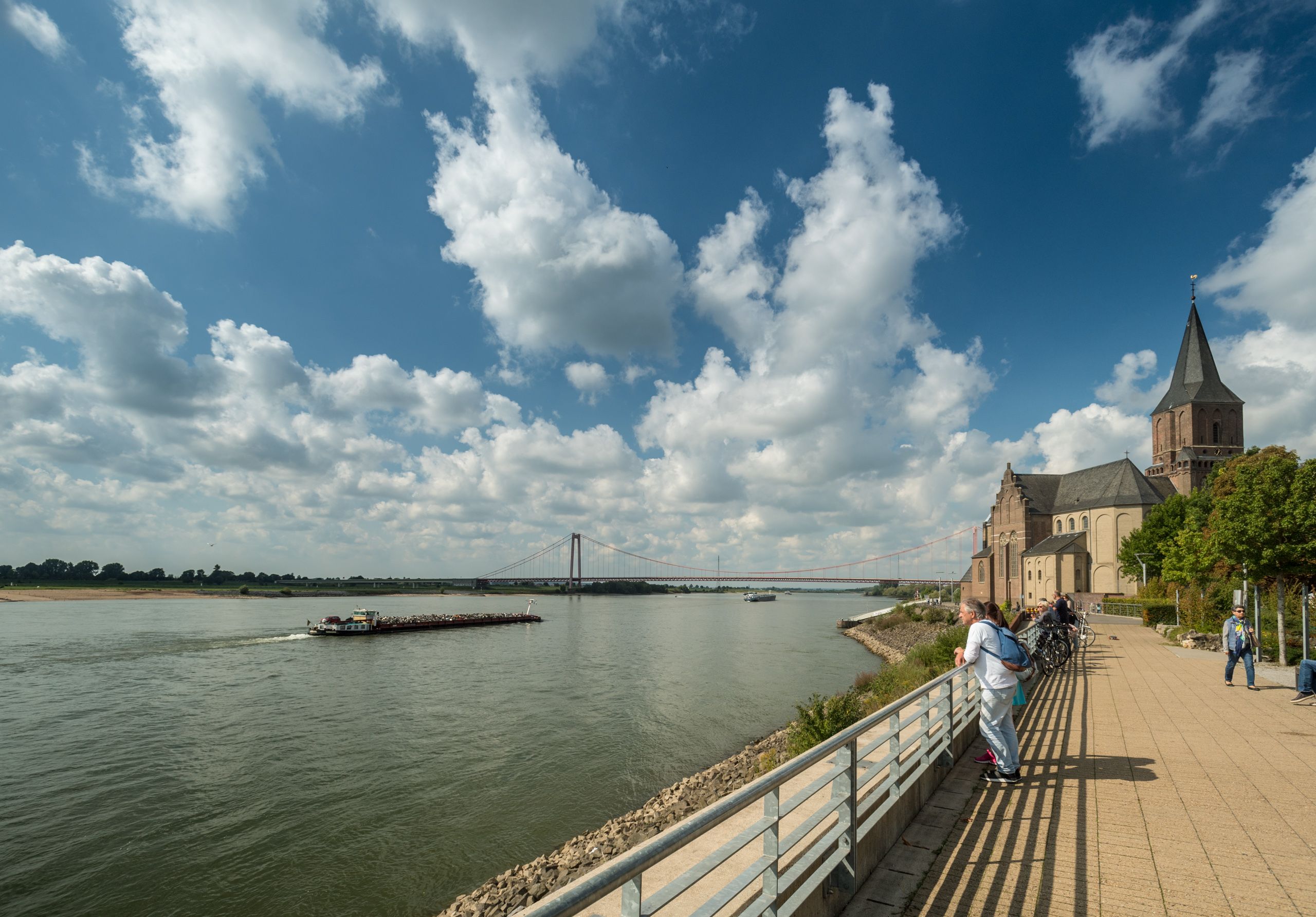 Ein Mann und eine Frau blicken auf den Rhein, im Hintergrund steht eine Kirche