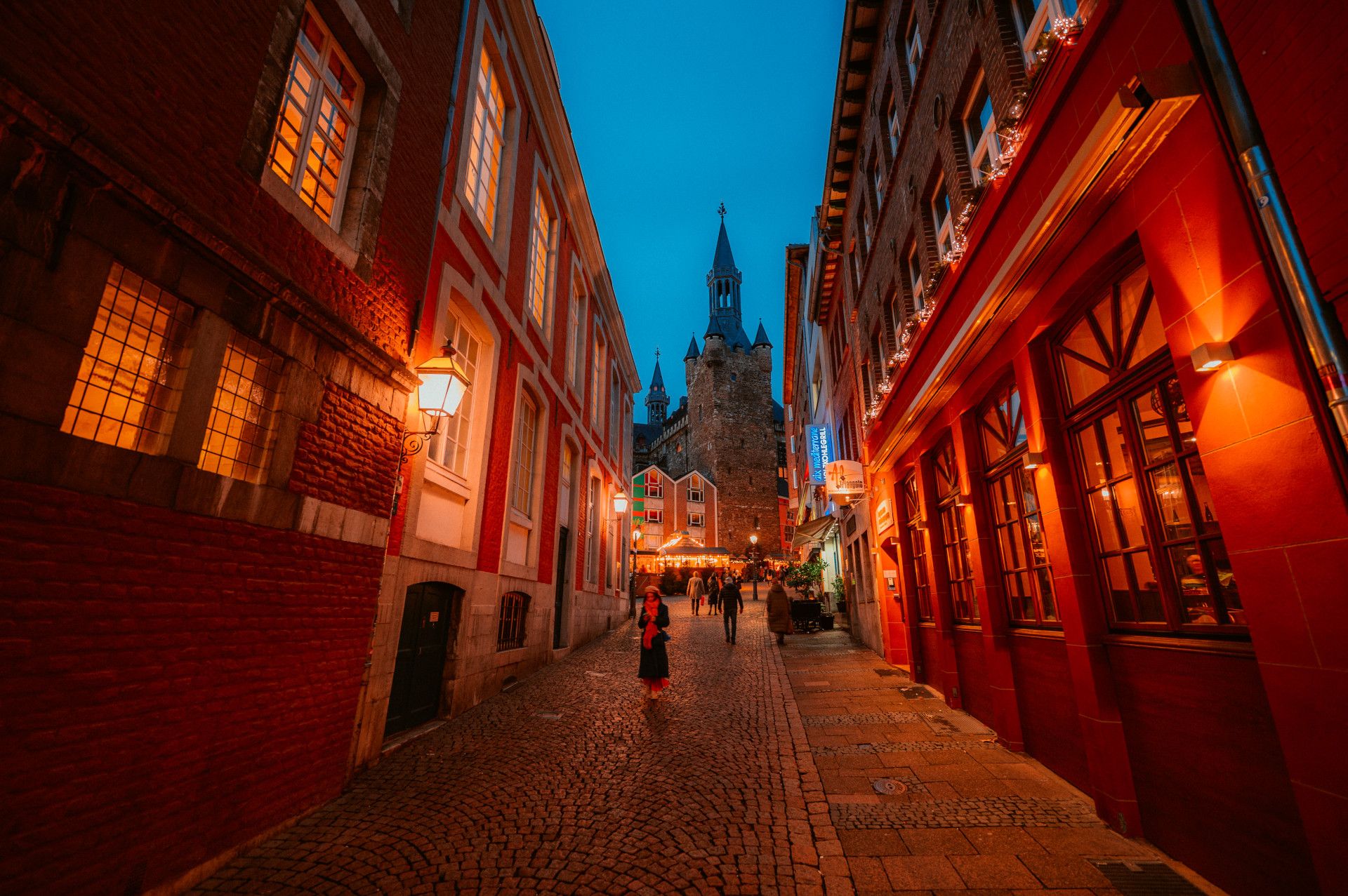An atmospheric alleyway in Aachen at dusk, lined with illuminated buildings. Aachen Cathedral can be seen in the background.