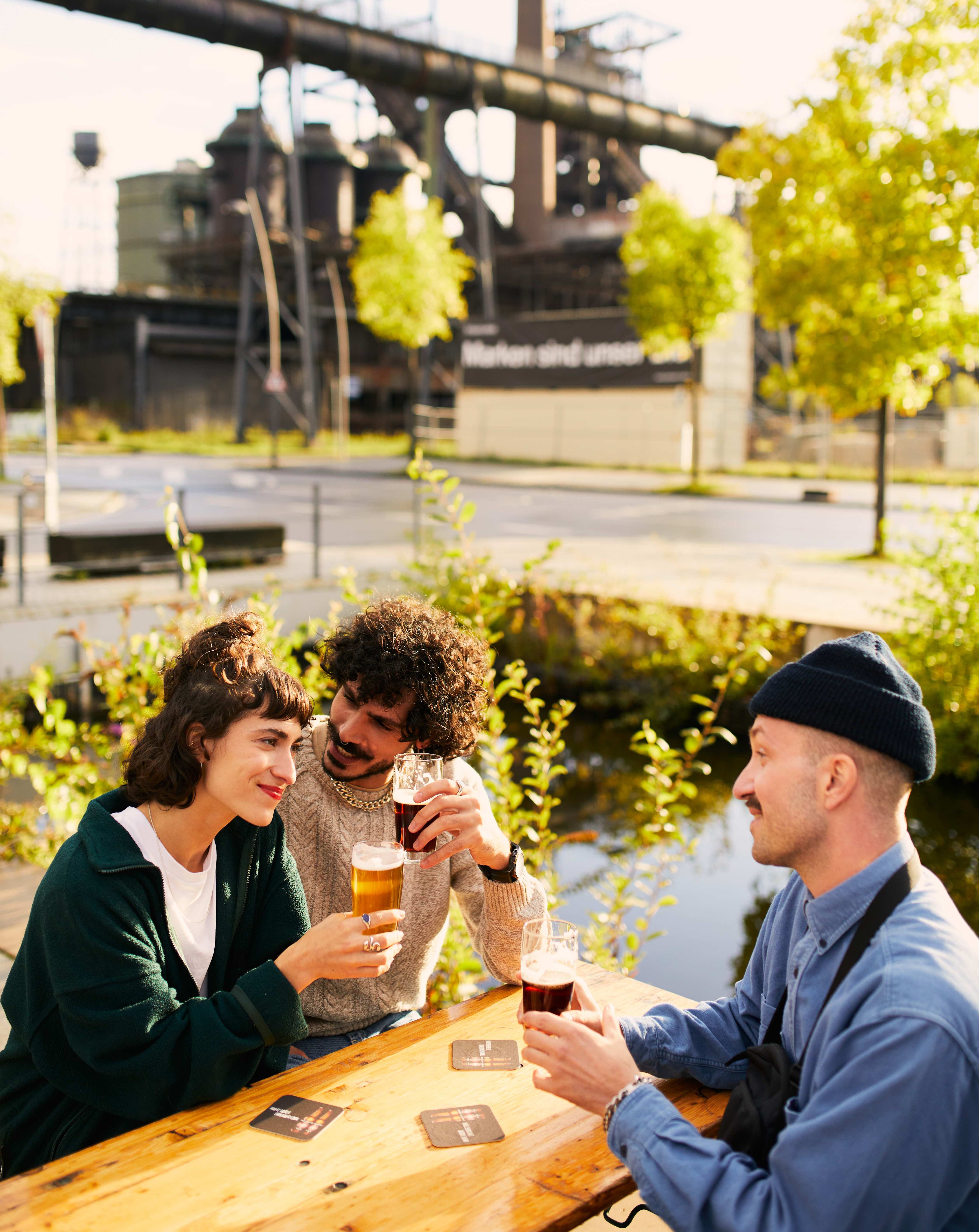Drei Menschen sitzen in der Sonne vor Industriekulisse mit Hochofen im Hintergrund und stoßen mit Biergläsern an