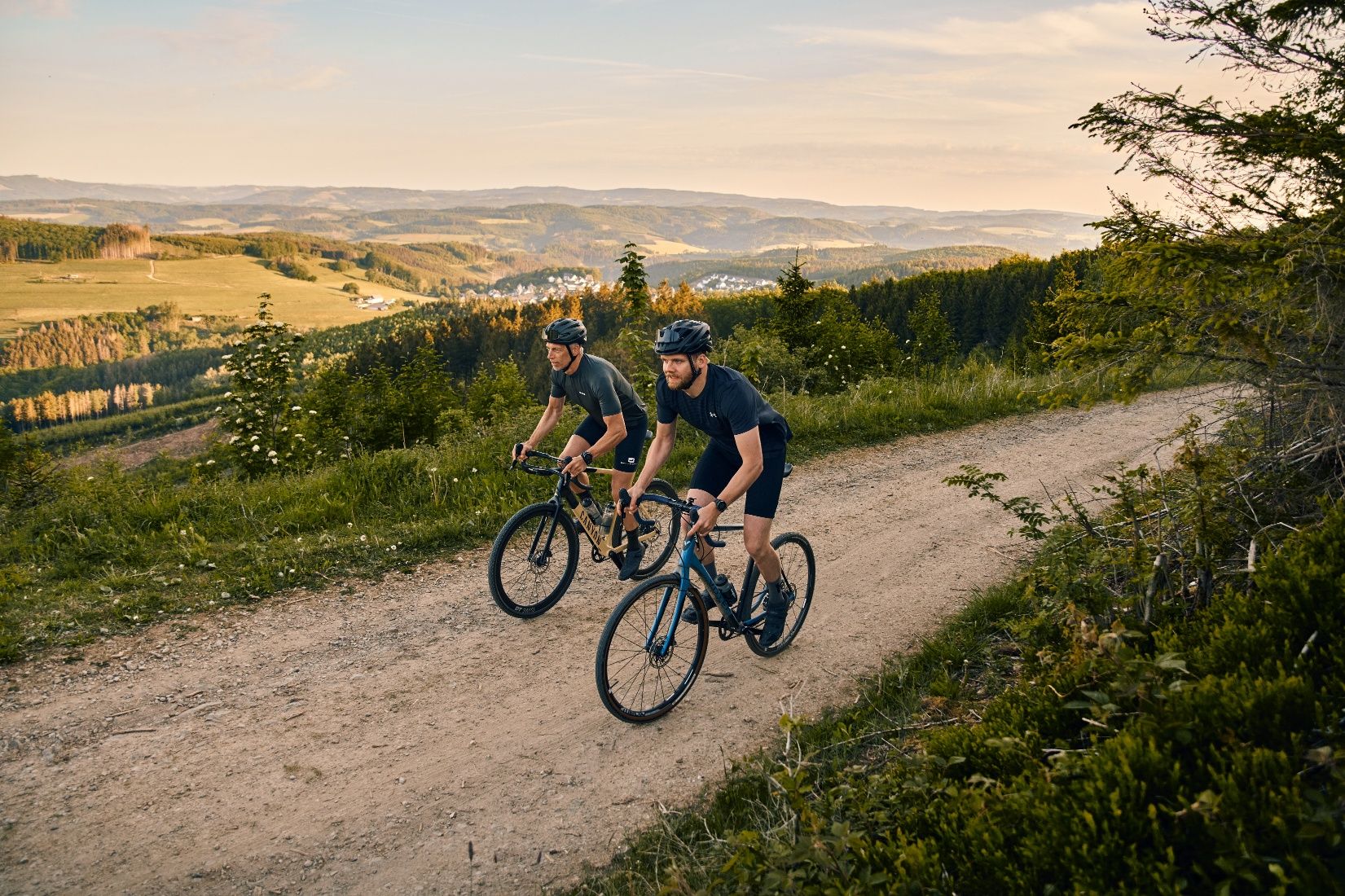 Two gravel bikers riding on a gravel path, with a view of the countryside behind them