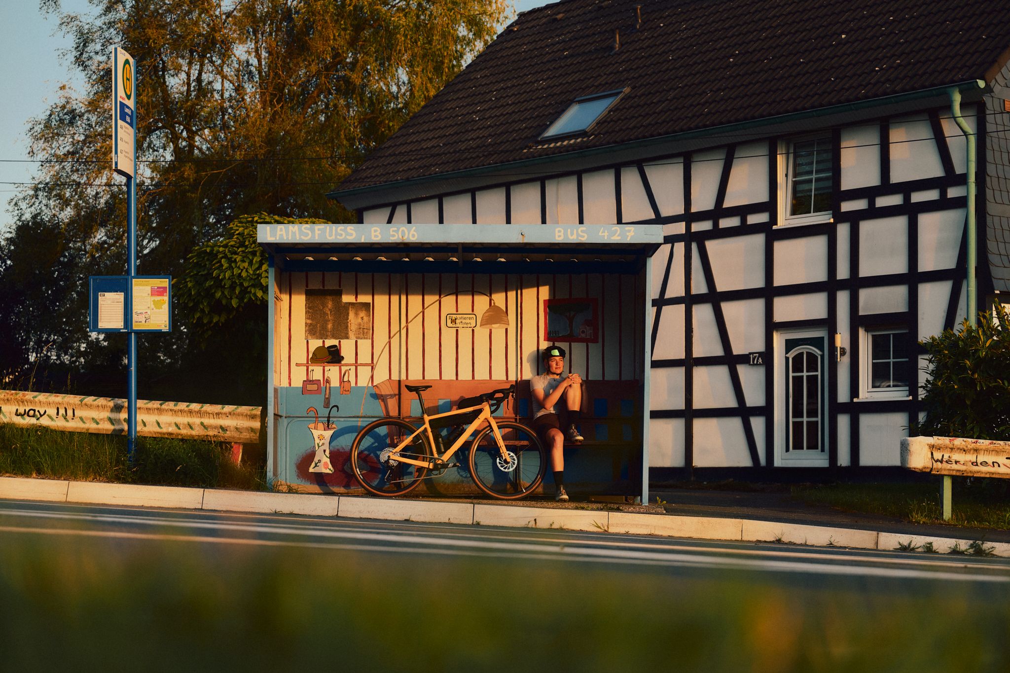 A person is sitting with a bicycle at a bus stop in front of a half-timbered house in the evening light.