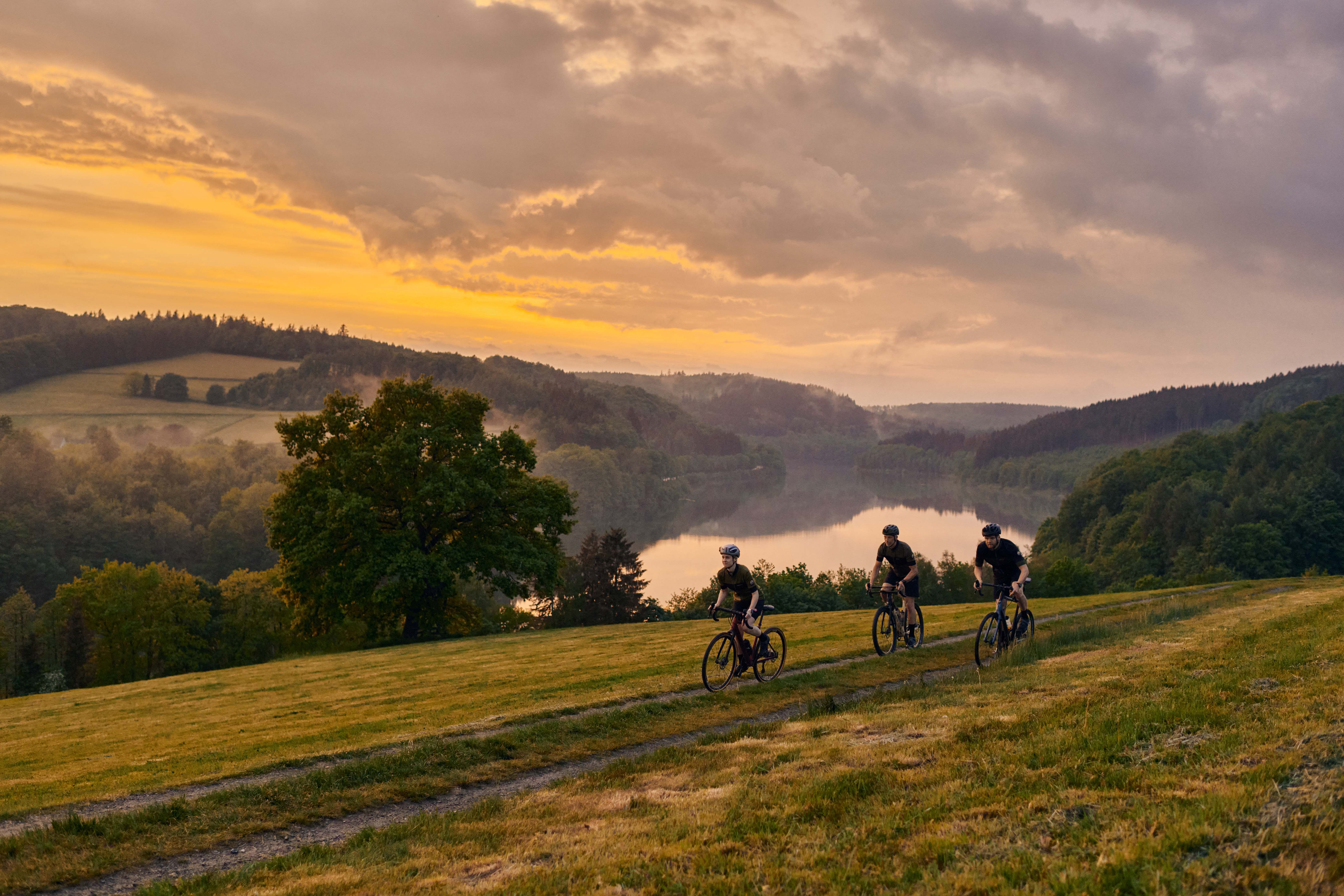 Three Persines ride gravel bikes through the hilly landscape with a lake in the background in the evening light