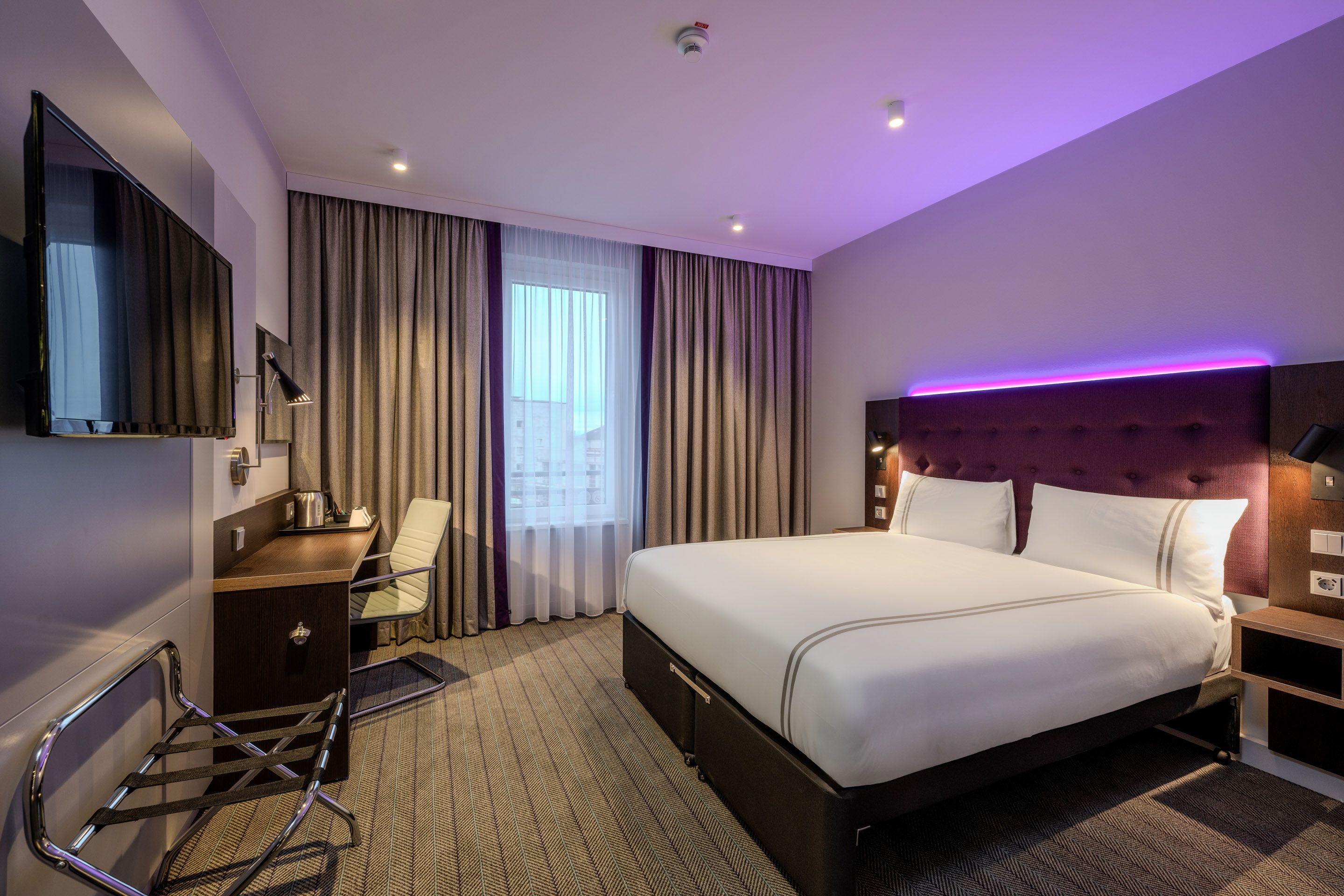 A modern hotel room with a large double bed, a desk with a chair, and a flat-screen TV mounted on the wall.