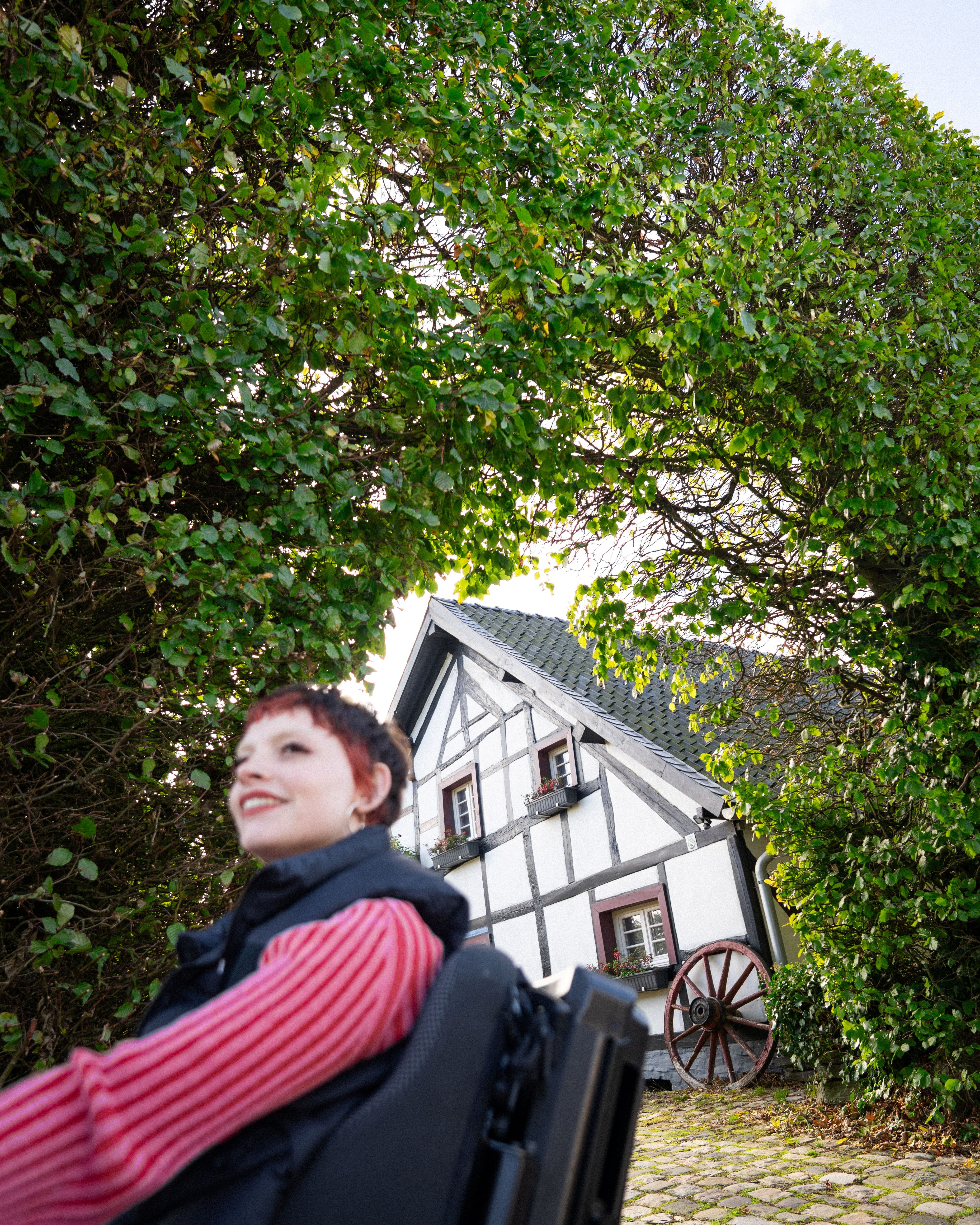 Wheelchair user pauses in front of a hedge with a view of a half-timbered house in Monschau-Höfen