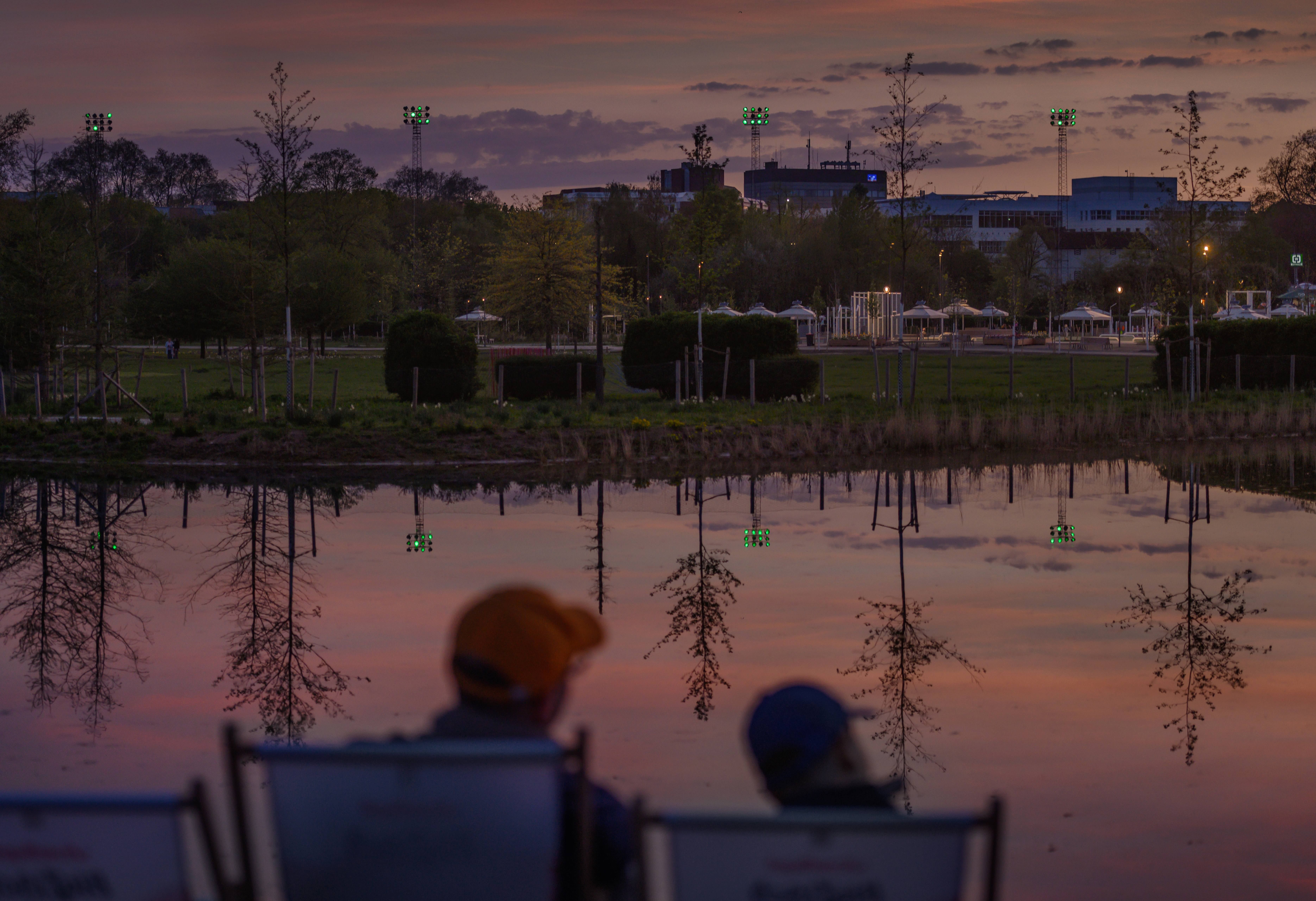Two people sit in deckchairs in front of a lake at sunset on the grounds of the Neuss 2026 State Garden Show.