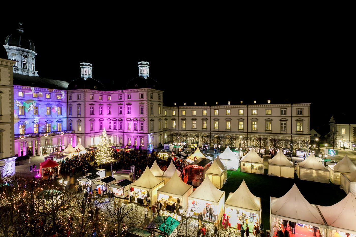 The courtyard of Bensberg Castle is illuminated with colorful lights, surrounded by white tents and a large Christmas tree.