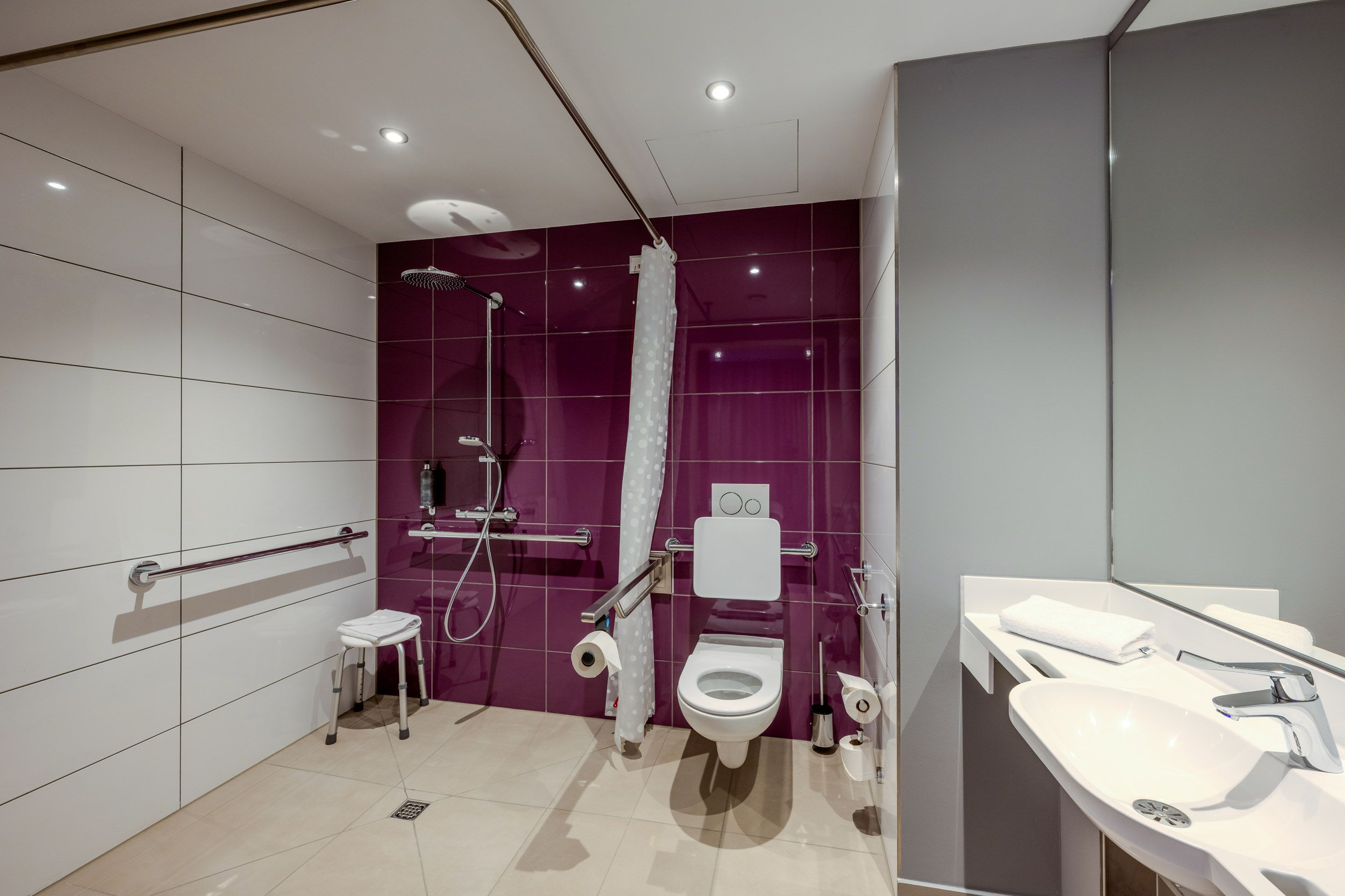 Modern, accessible bathroom with purple tiles, a shower, a toilet, and a sink. Grab bars and a shower stool are provided.