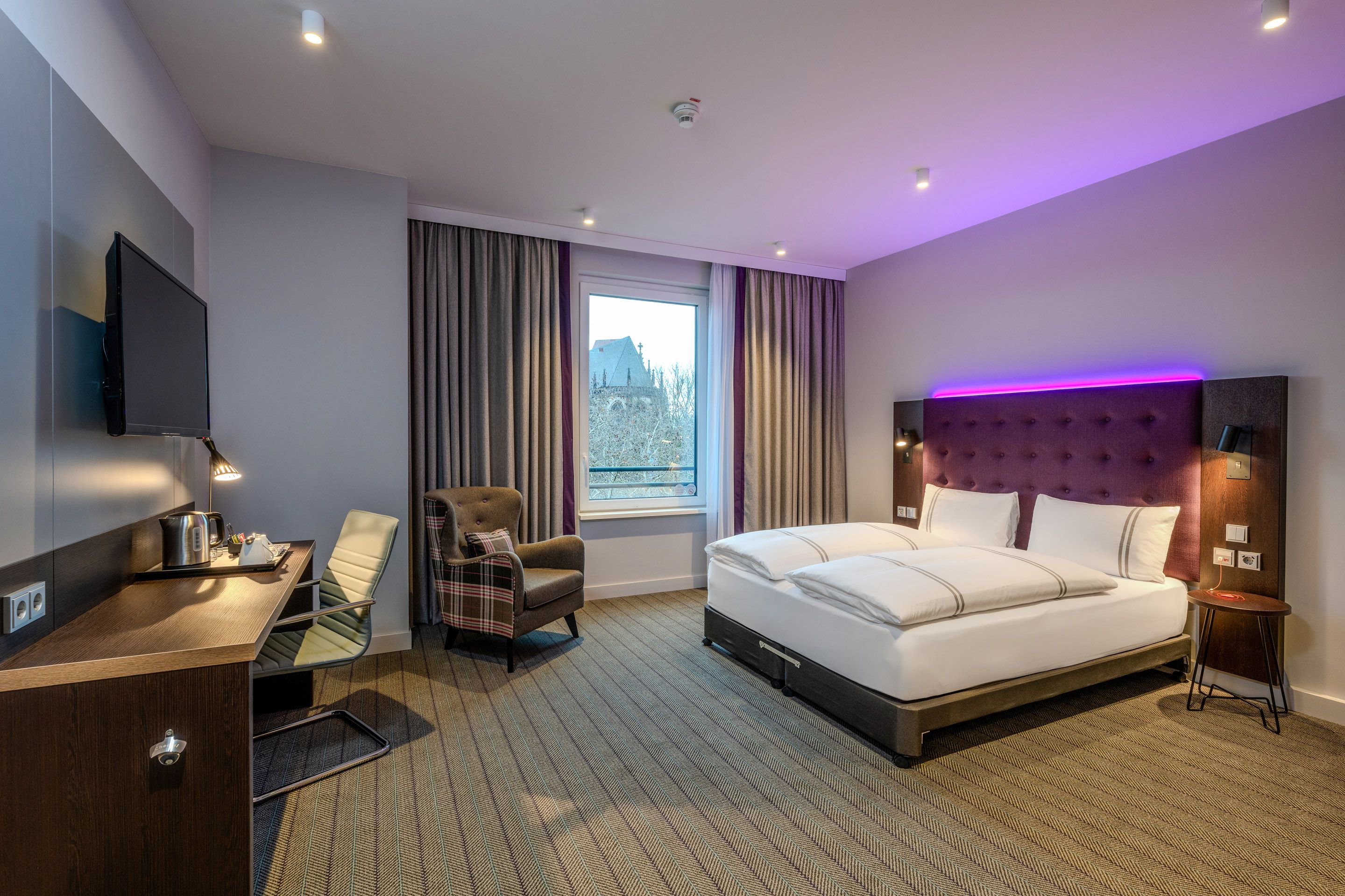 A modern hotel room with a large double bed, a desk with a chair, an armchair, and a window overlooking a building outside.