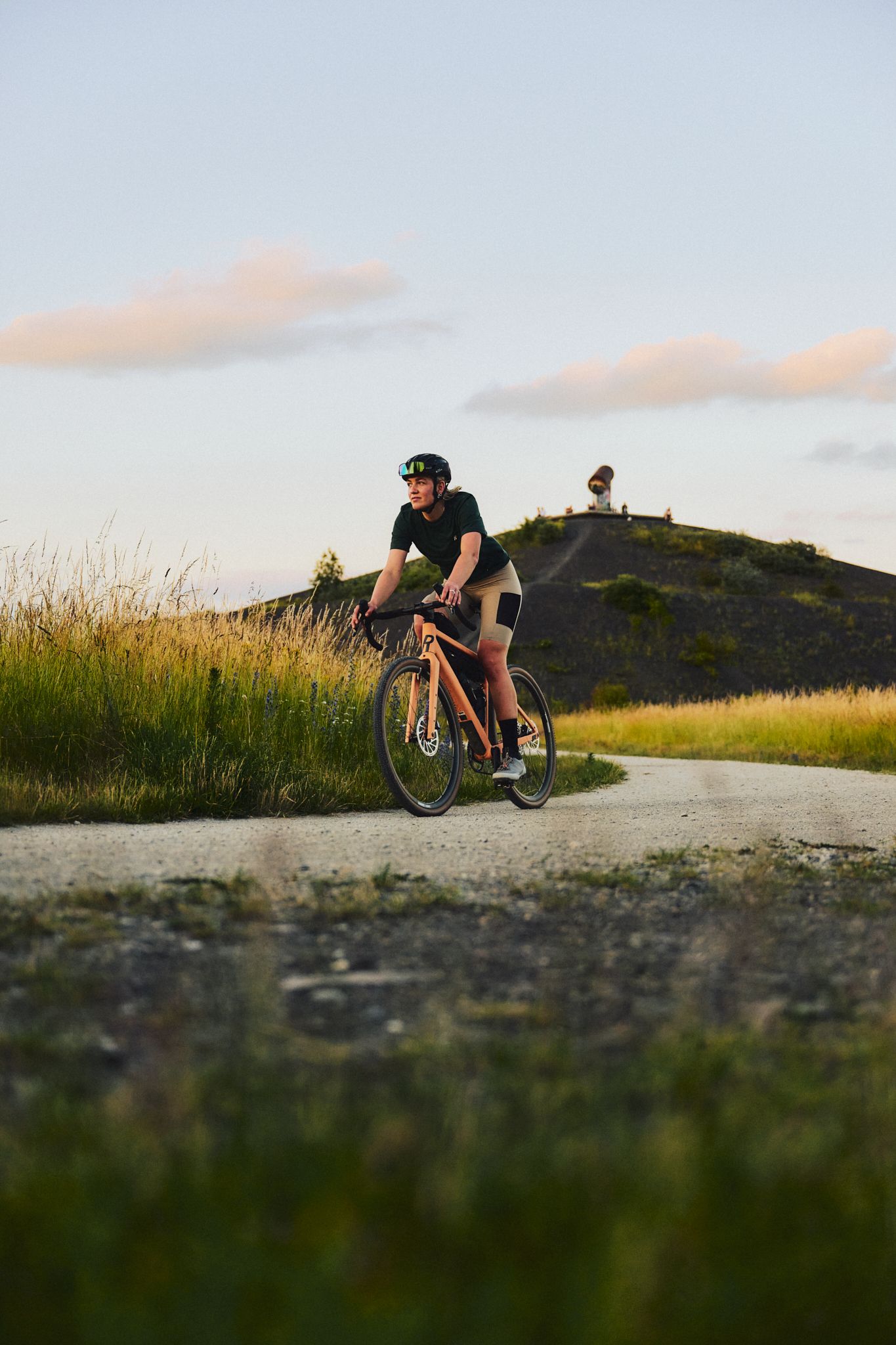 A woman is riding a gravel bike on a gravel path in the Ruhr region. The Rungenberg slag heap can be seen in the background.