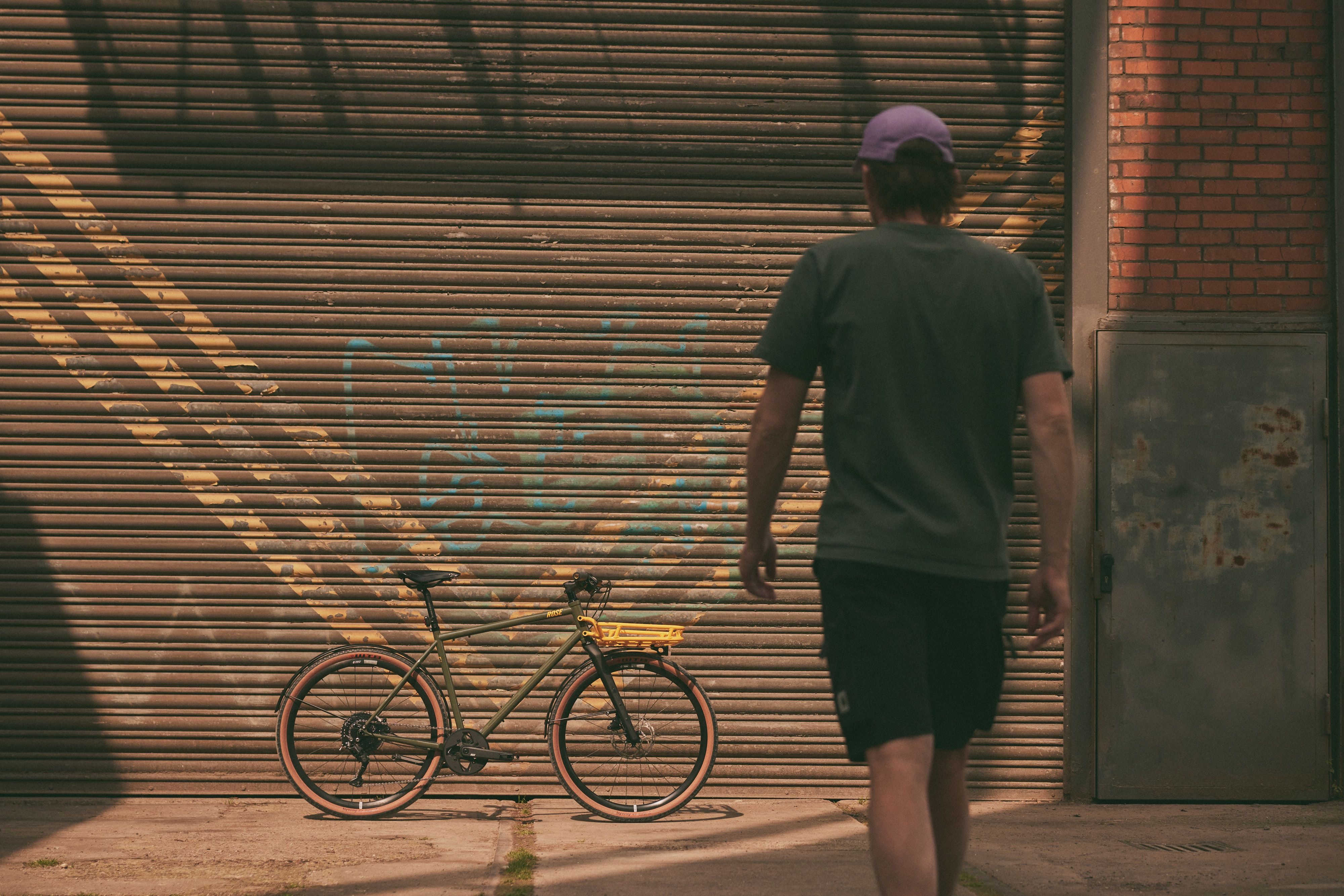 A gravel bike stands on an abandoned industrial site with old pipes and brick buildings in the background.