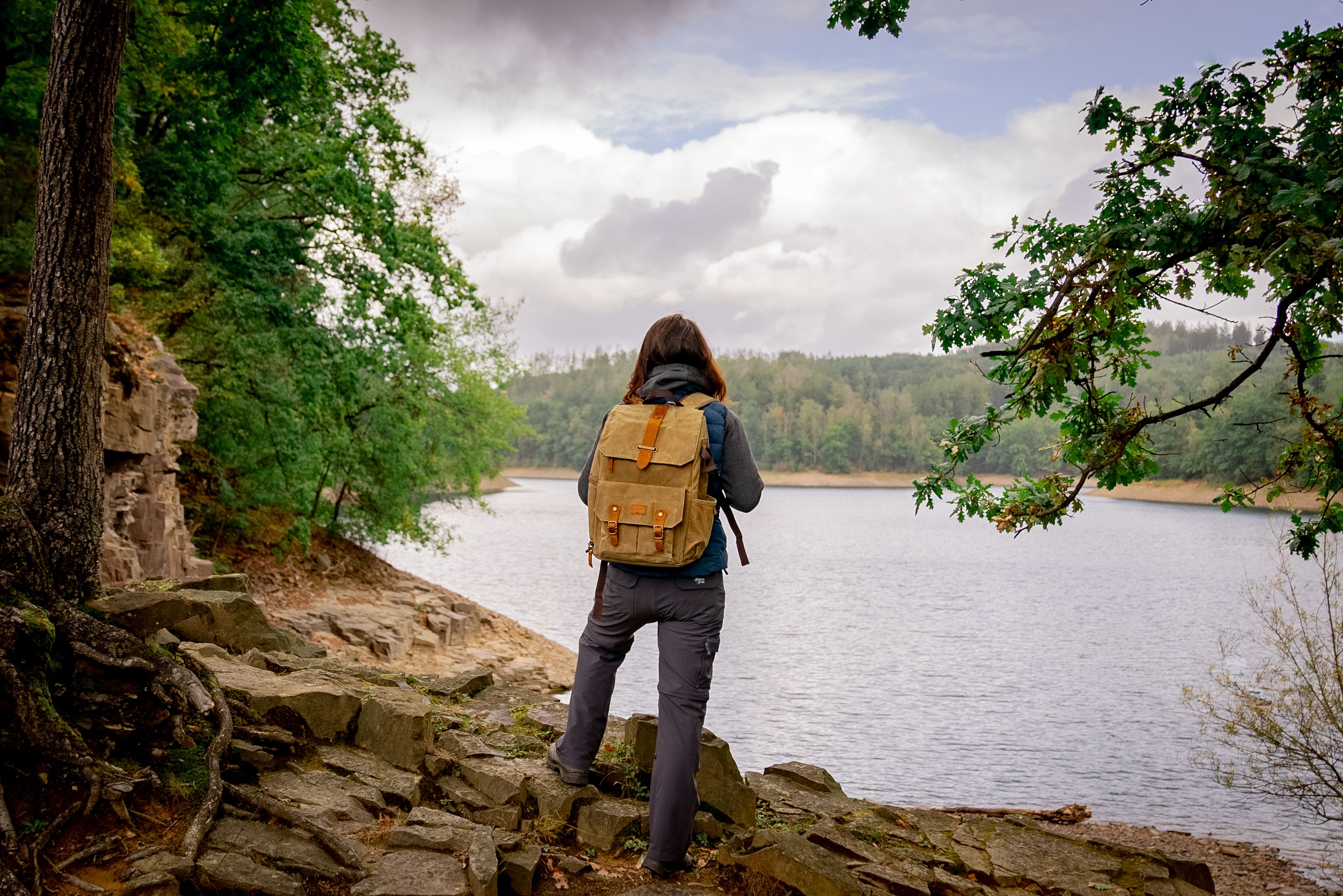 A person with a backpack is standing on a rocky bank, looking out at the Aggertal Dam, which is surrounded by trees.