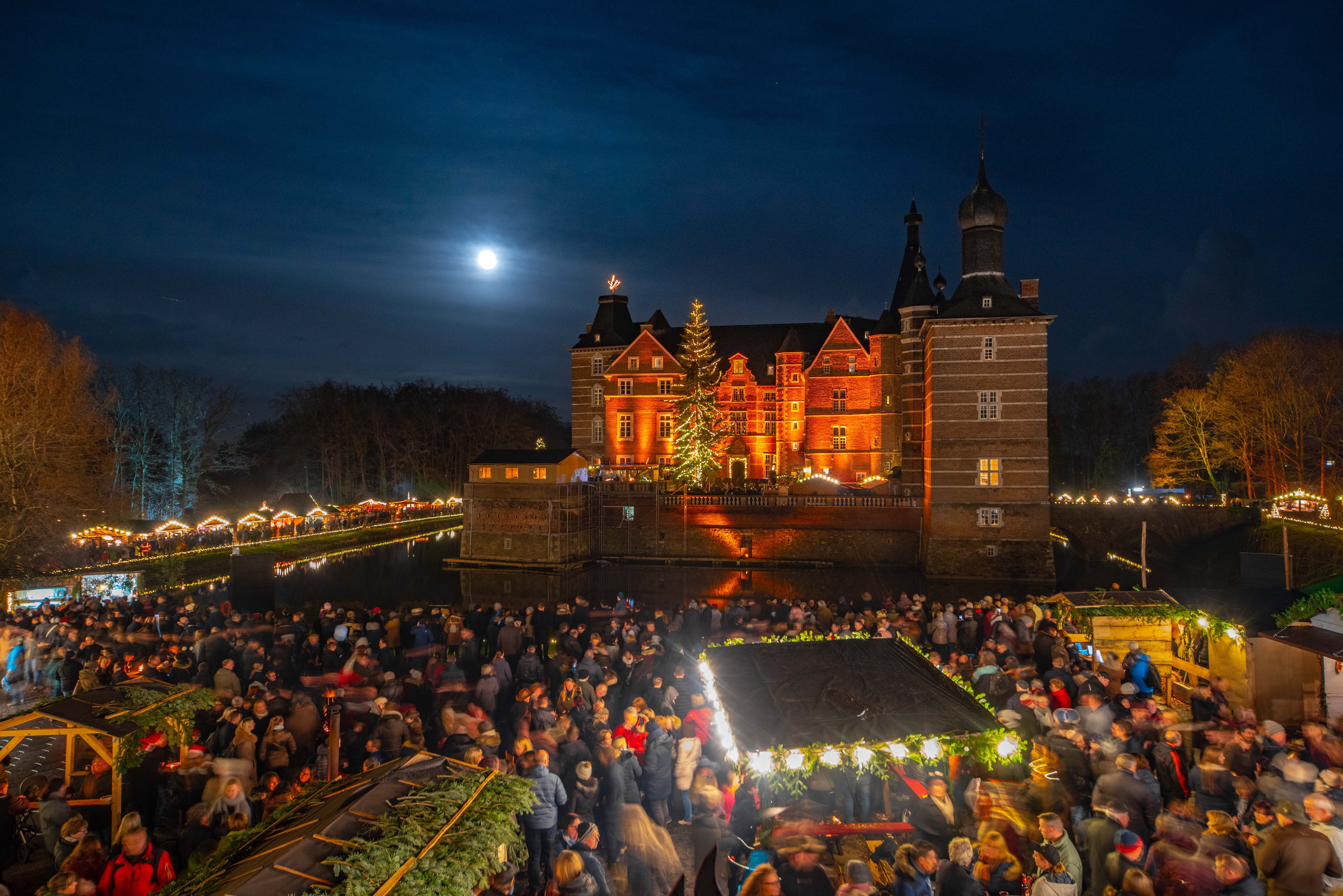 Merode Castle at night, festively illuminated with a Christmas market and numerous visitors. The full moon shines in the sky.