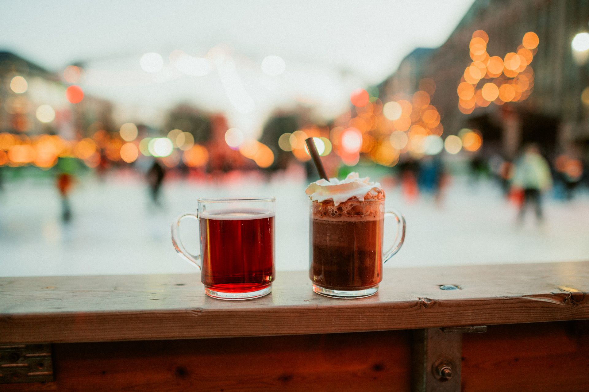 Two glasses of mulled wine and hot chocolate on a wooden table, with a blurred ice rink with lights in the background.