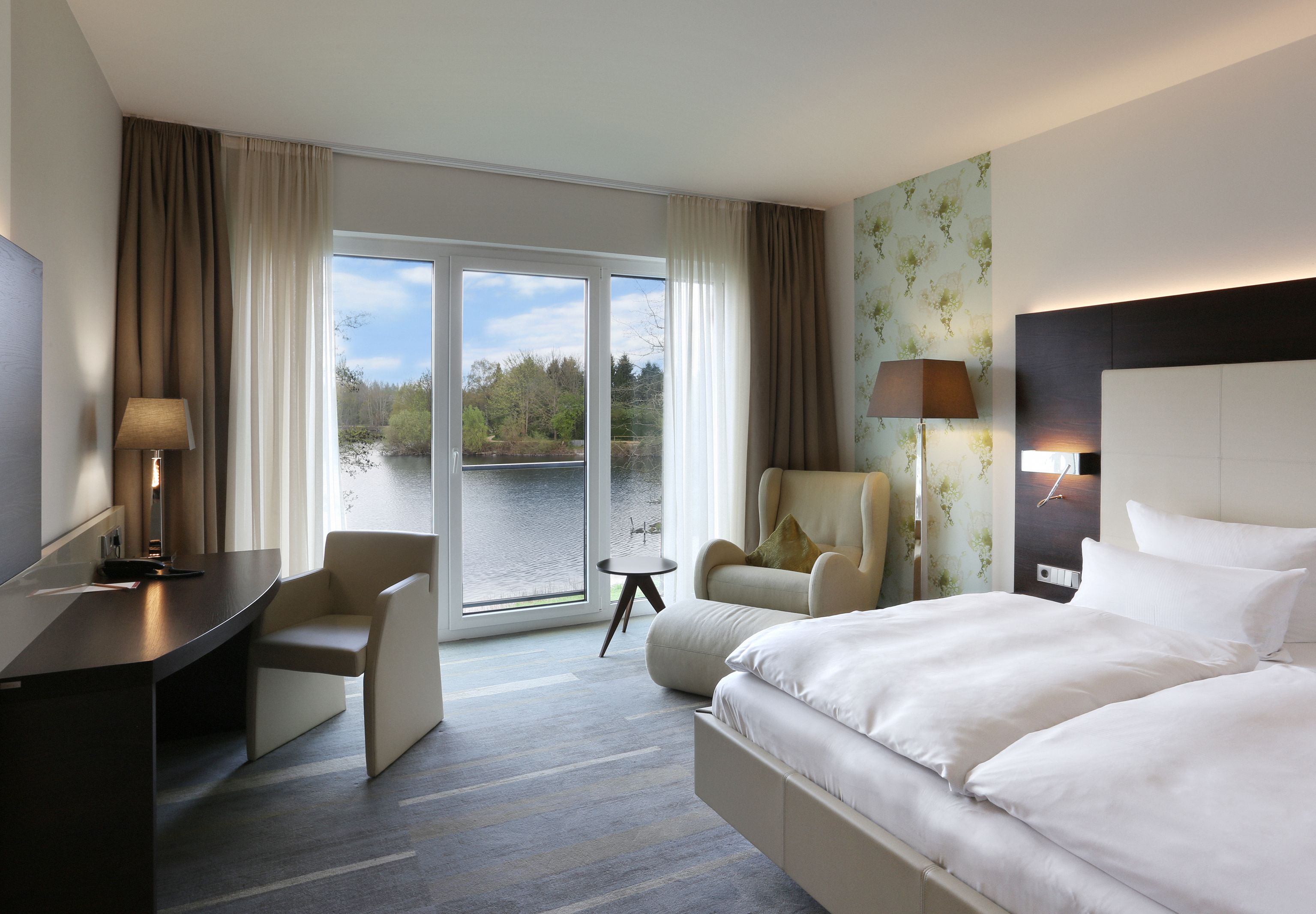 Modern hotel room with double bed, armchair and desk, view of a lake through large windows in Geldern
