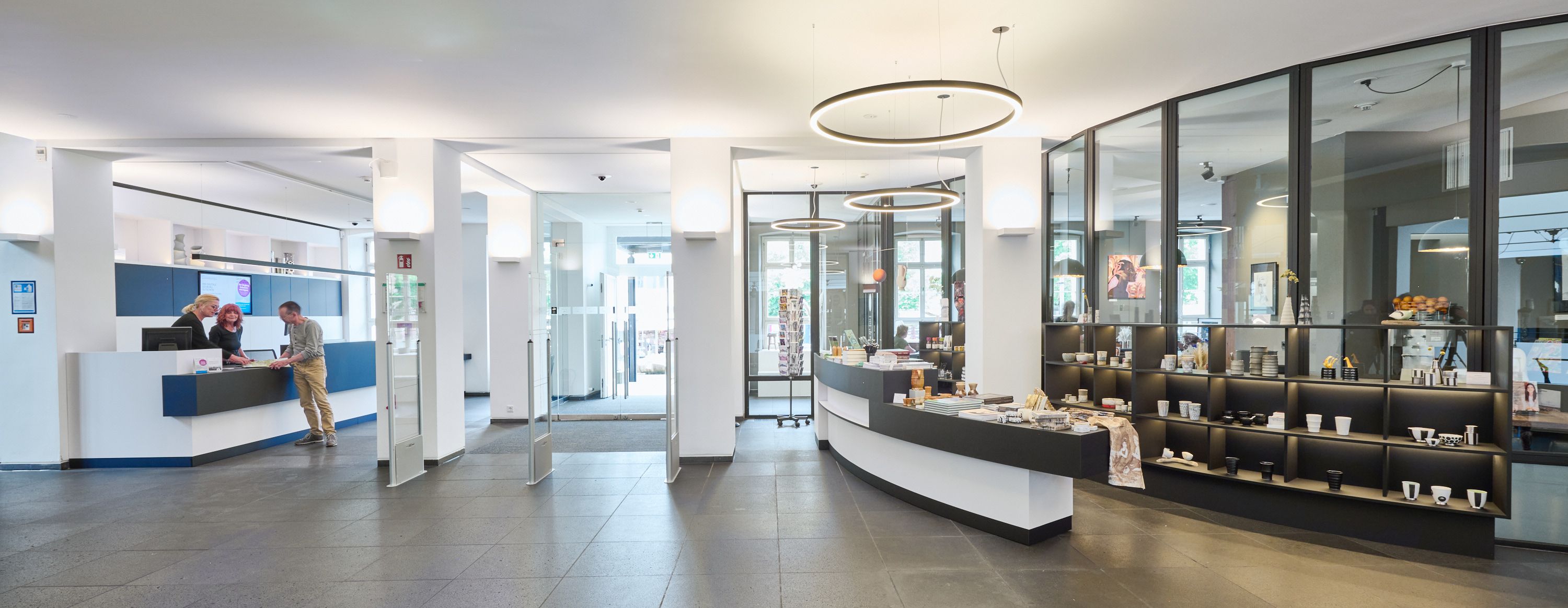 A bright interior of the Siegburg Tourist Information Center, featuring a reception area, sales shelves, and three people at the counter.