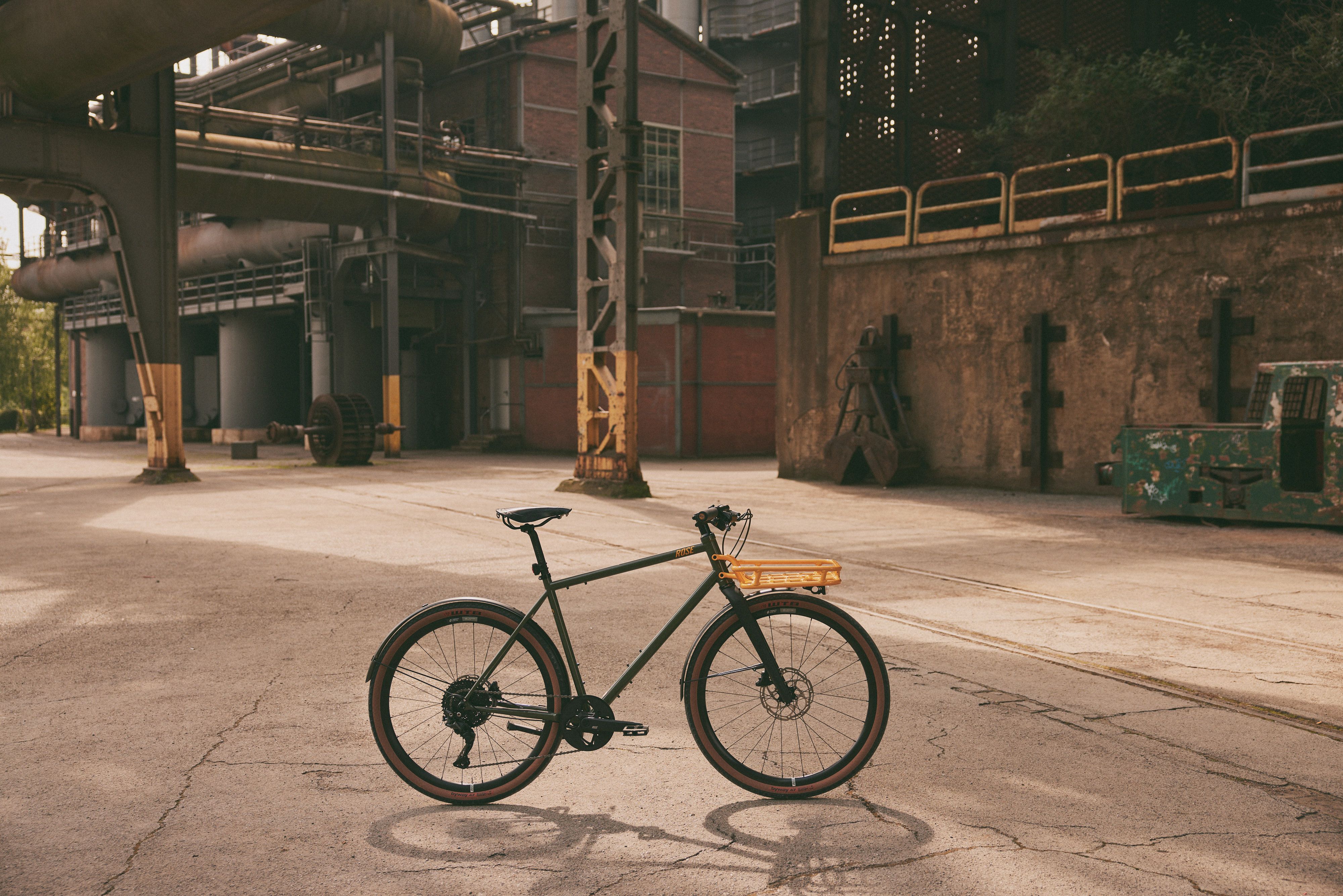 A gravel bike stands on an abandoned industrial site with old pipes and brick buildings in the background.