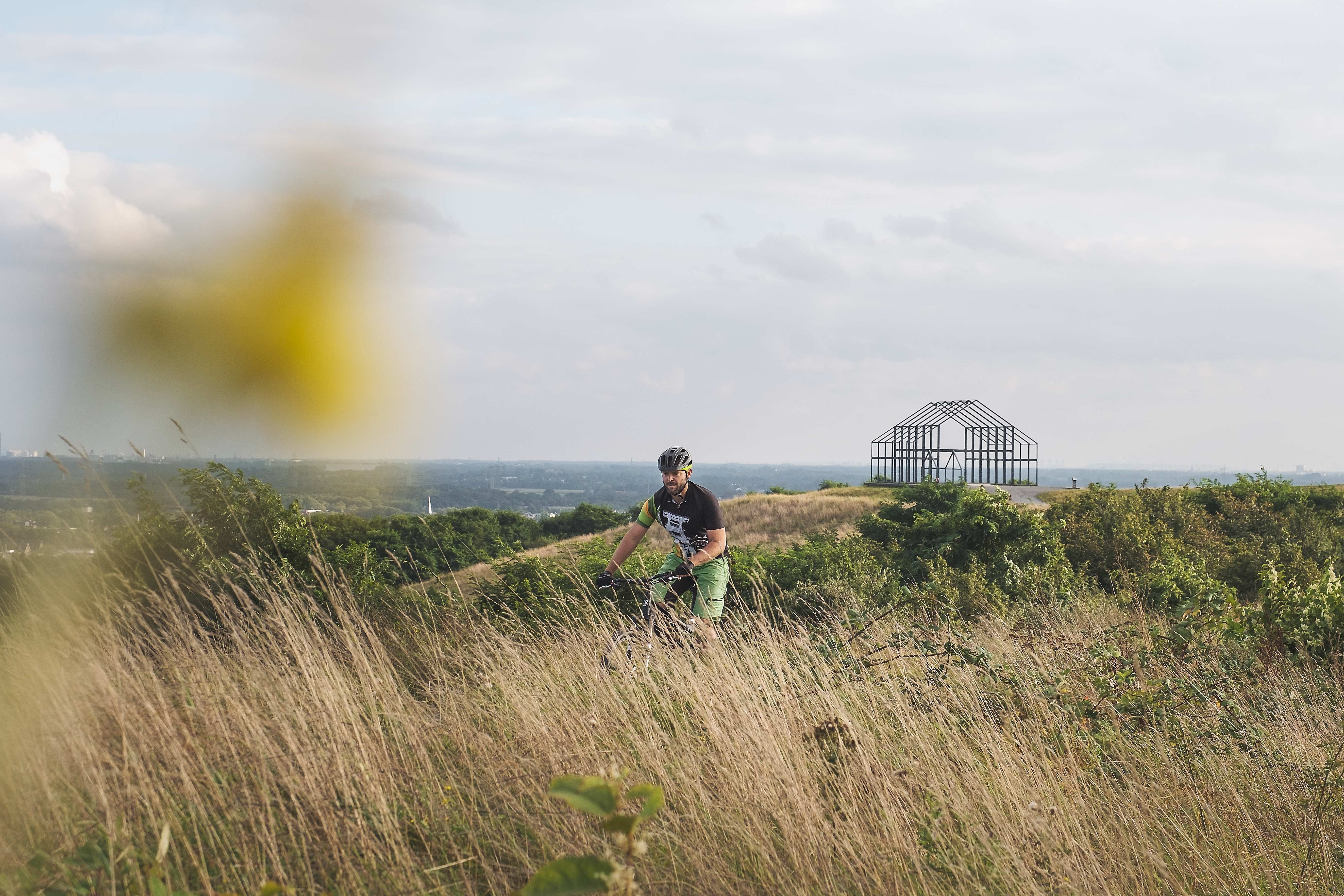 Radler unterwegs in hügeliger Haldenlandschaft mit Skulptur "Hallenhaus" im Hintergrund