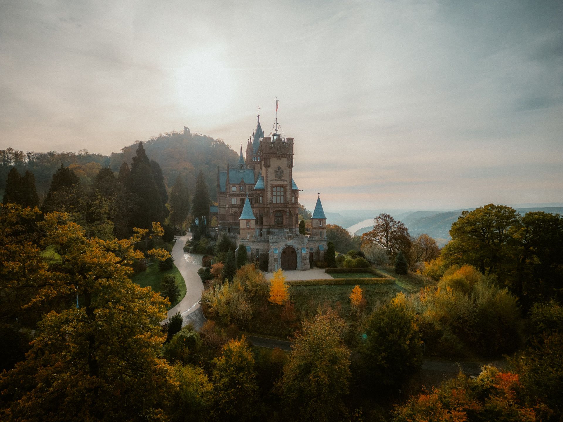 Drachenburg Castle, surrounded by colorful foliage and rolling hills at sunset. The Rhine flows in the background
