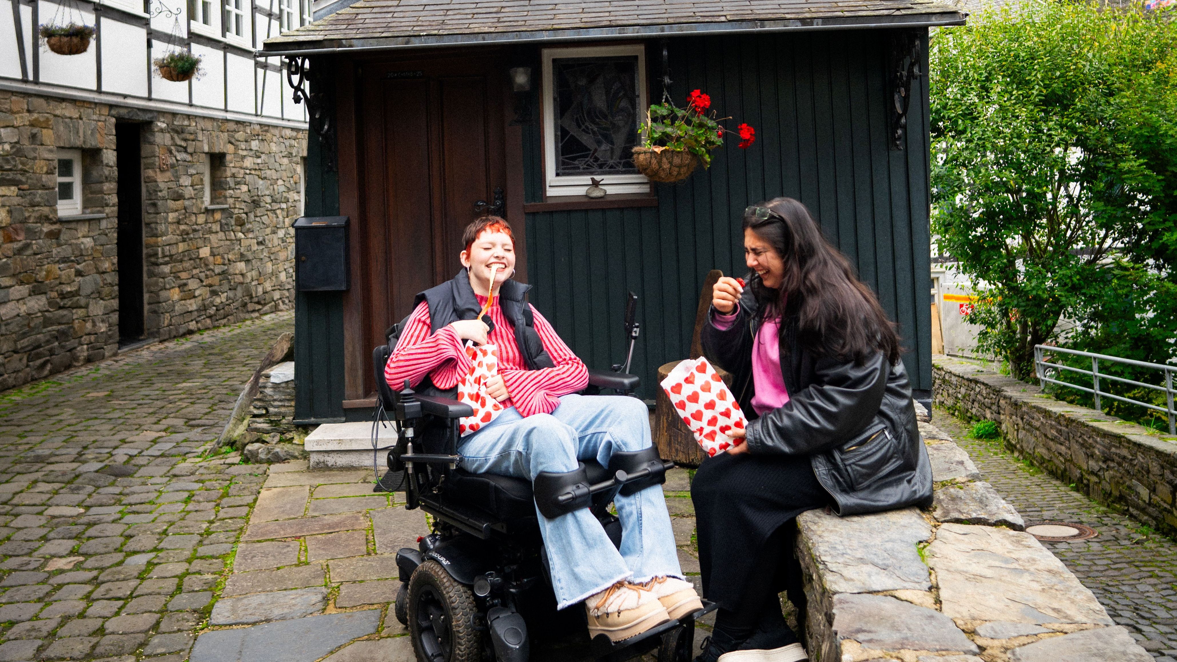 Two laughing friends enjoy sweets from bags in front of a half-timbered house in Monschau
