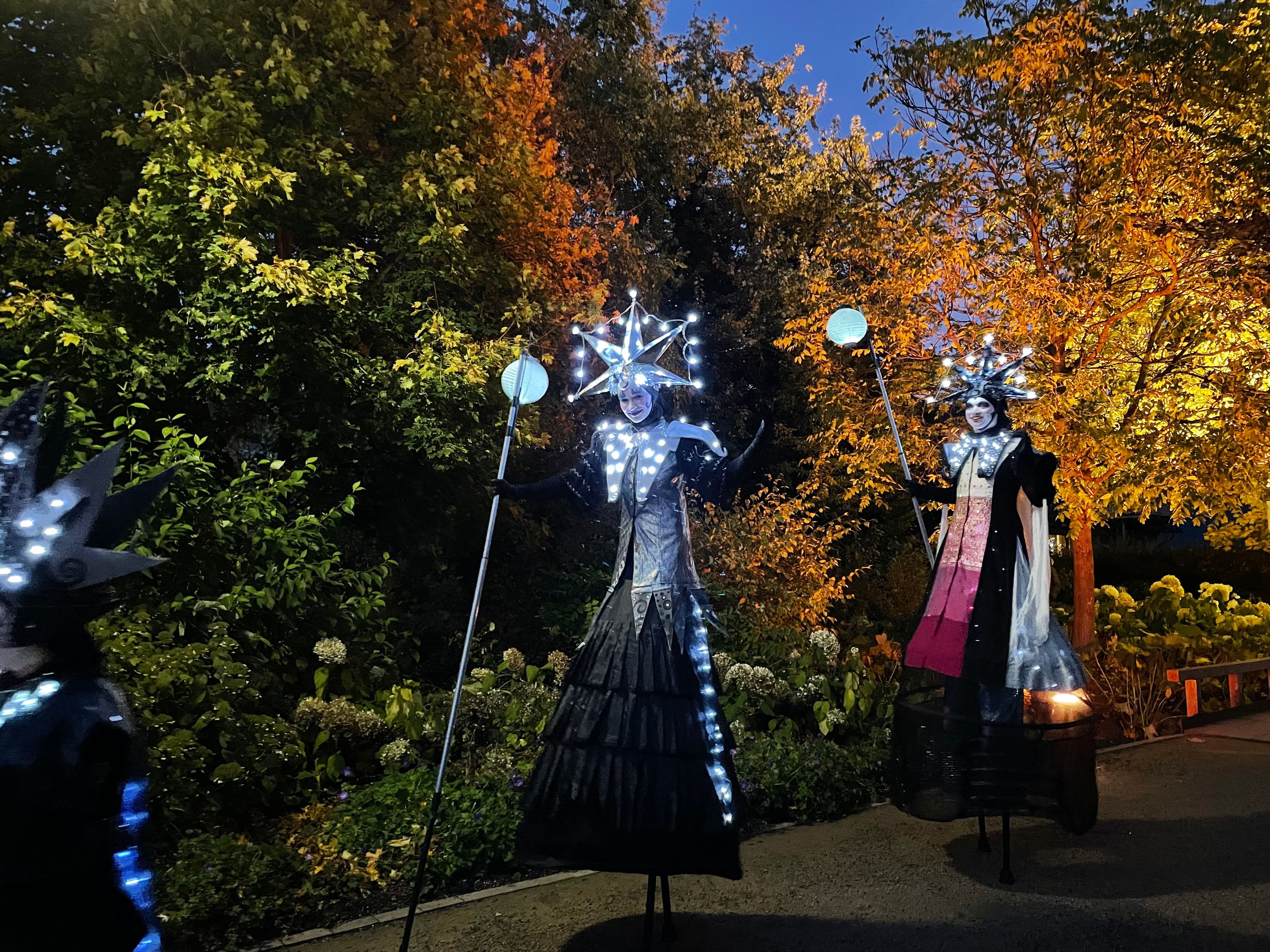 Two artists on stilts wear glowing costumes and hats in Lennestadt at night.