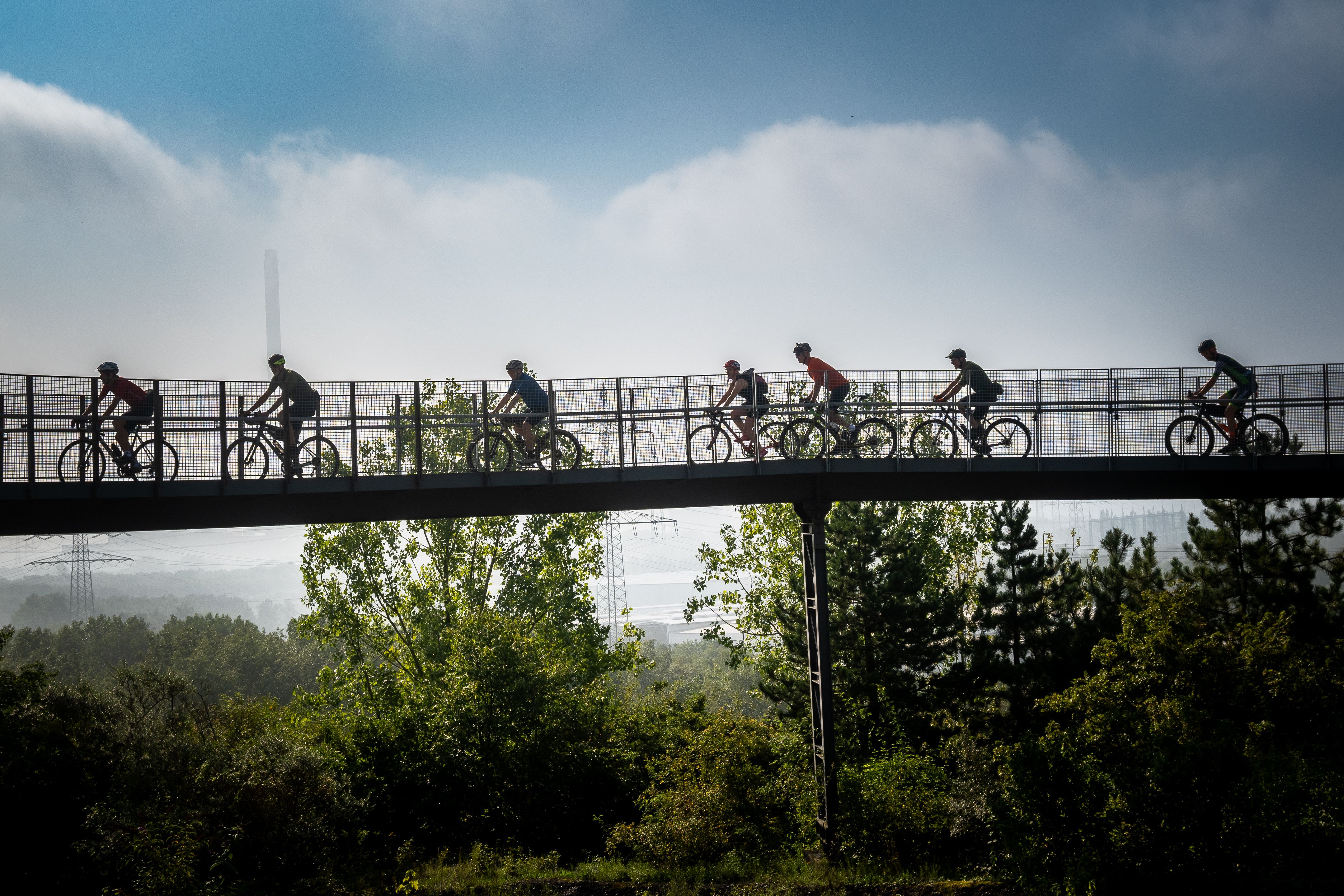 Silhouette of cyclists on a bridge, surrounded by trees and cloudy sky in the background.