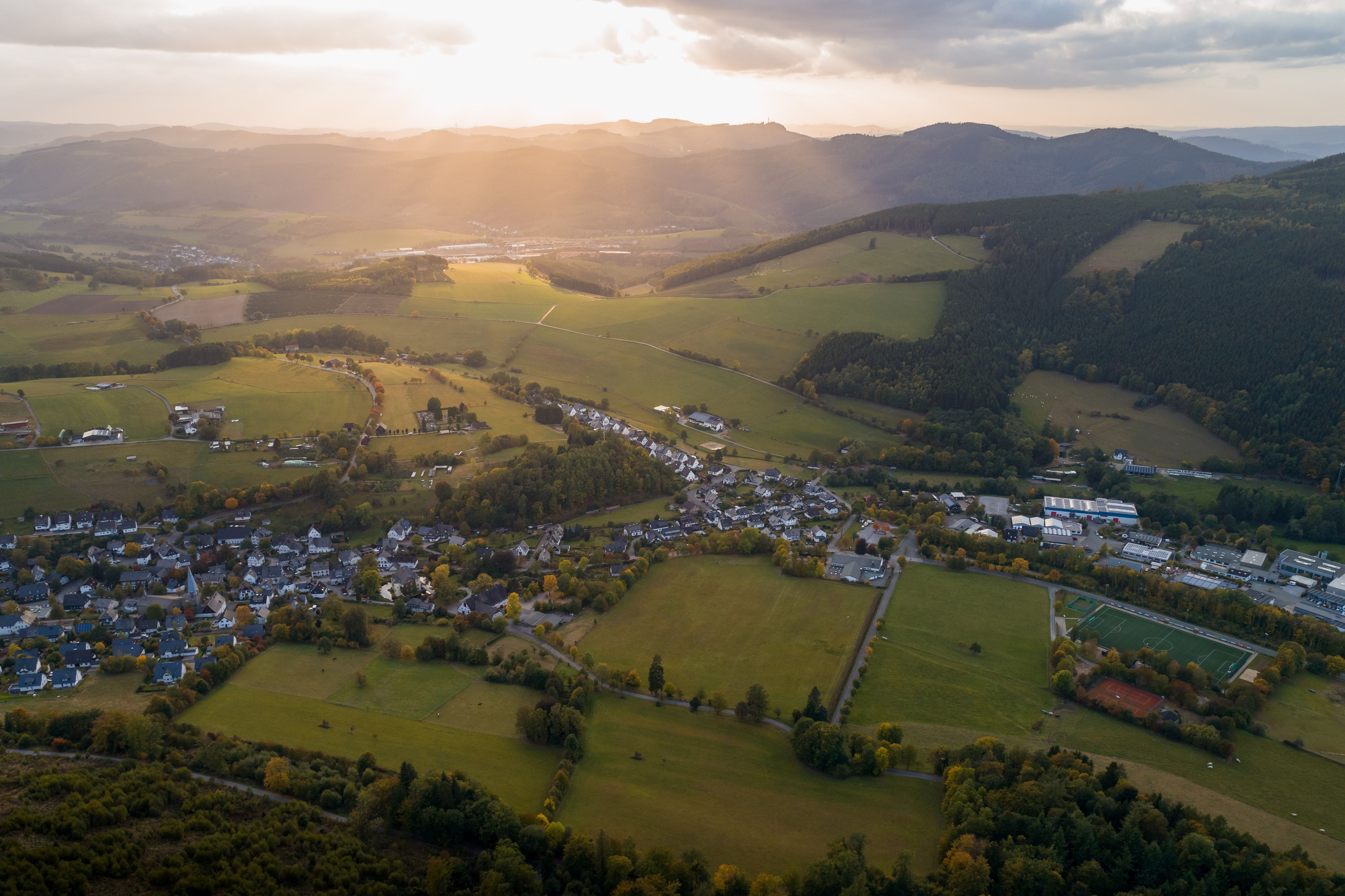 Aerial view of a rural area at sunset. Fields, forests and a village can be seen, surrounded by rolling hills and golden light.