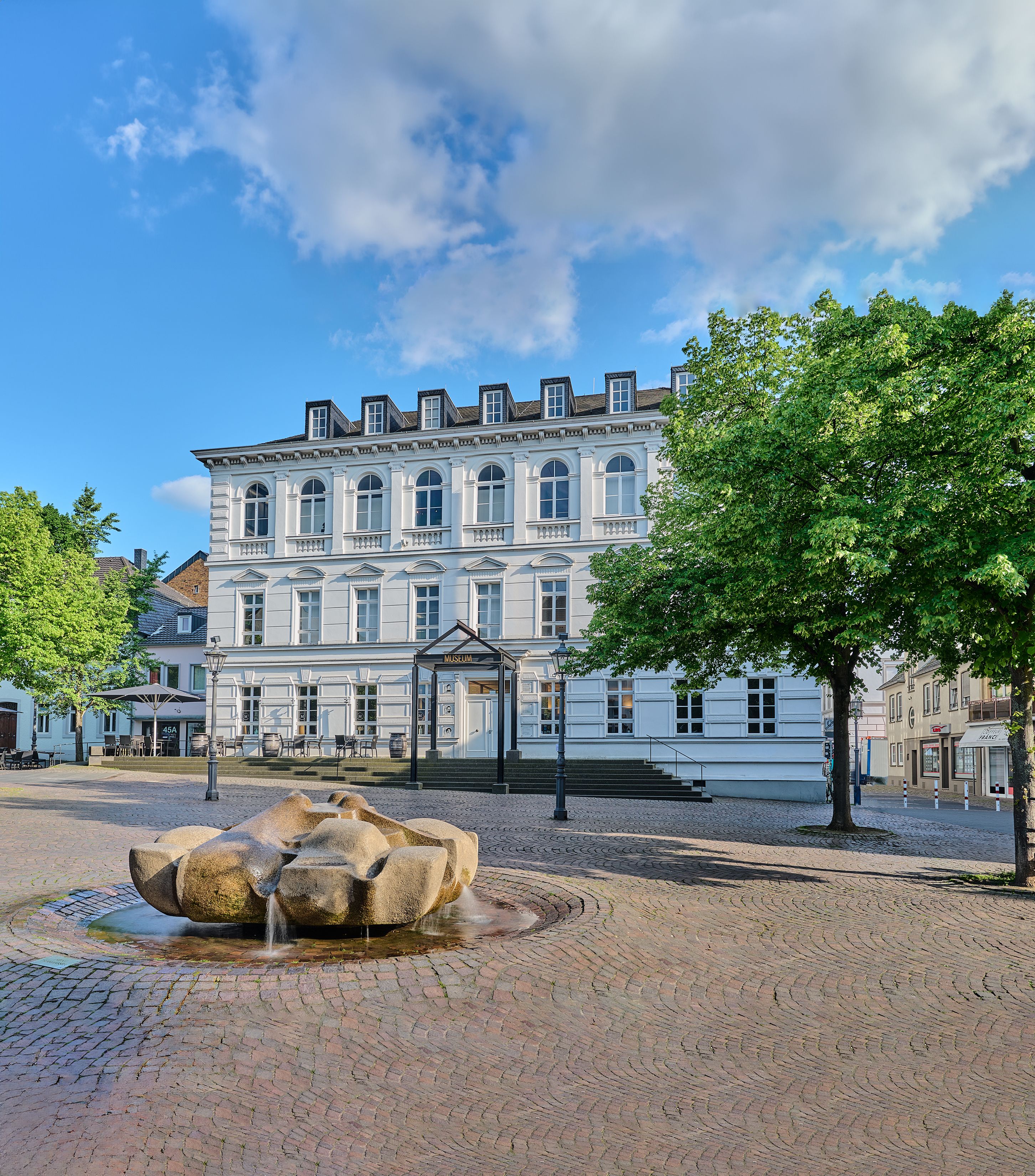 The Siegburg City Museum and Tourist Information Center are located on a sunny square with a fountain, trees, and a historic building.