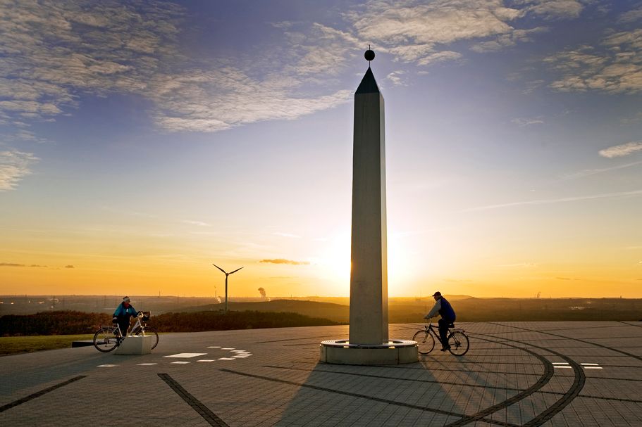 Zwei Radler am Abend auf Halde Hoheward fahren an Skulptur Sonnenuhr