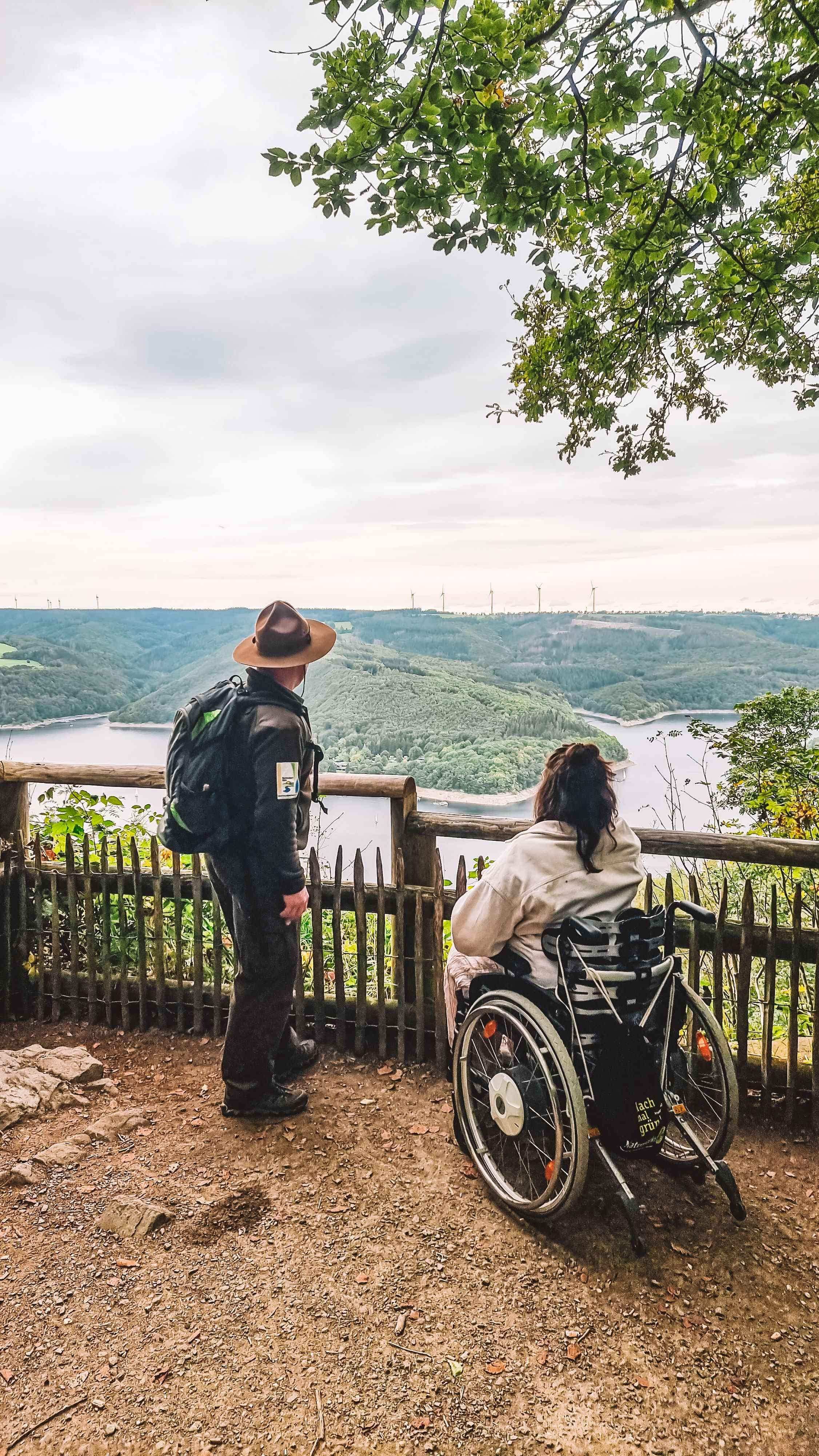 Ranger und Rollstuhlfahrerin blicken von Aussichtspunkt auf See und Wald