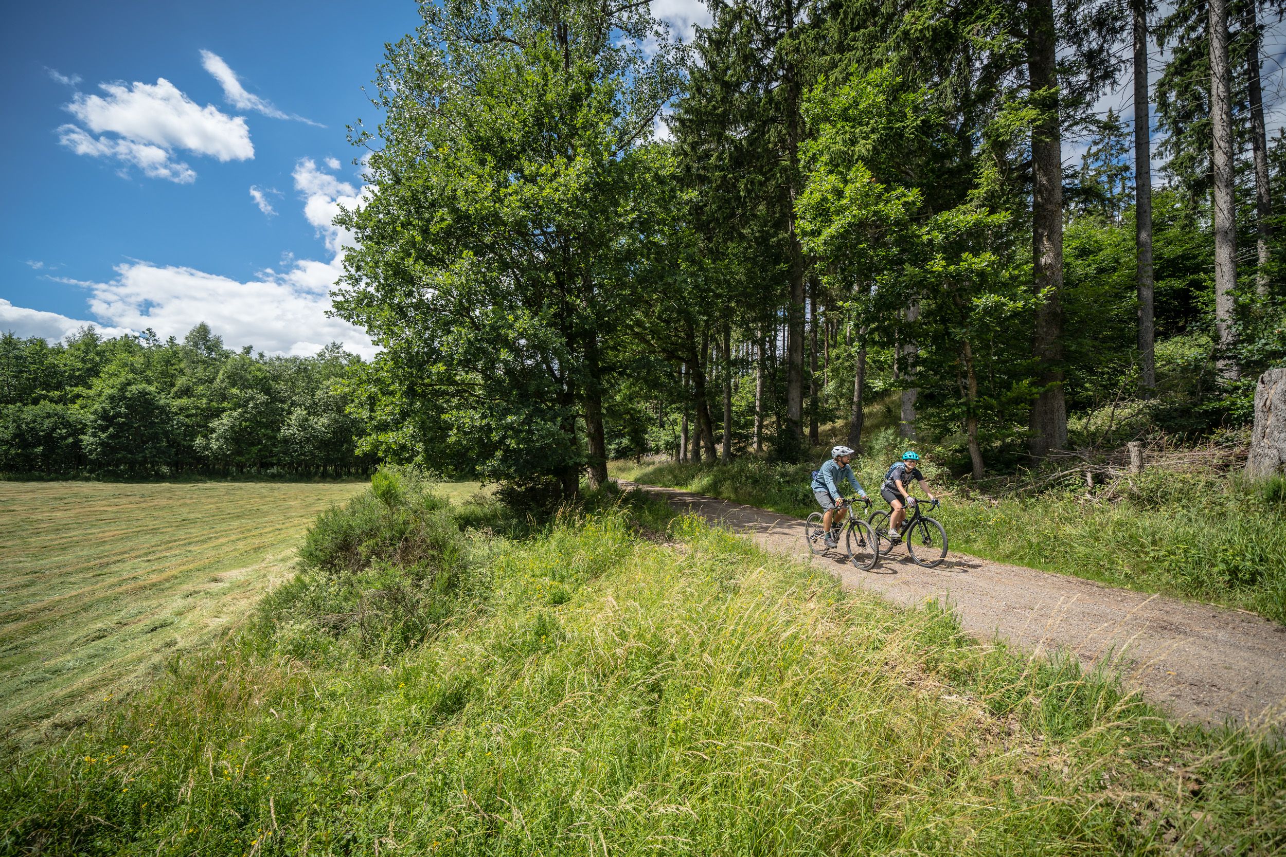 Two cyclists ride along a gravel path through a green forest in the Eifel region.